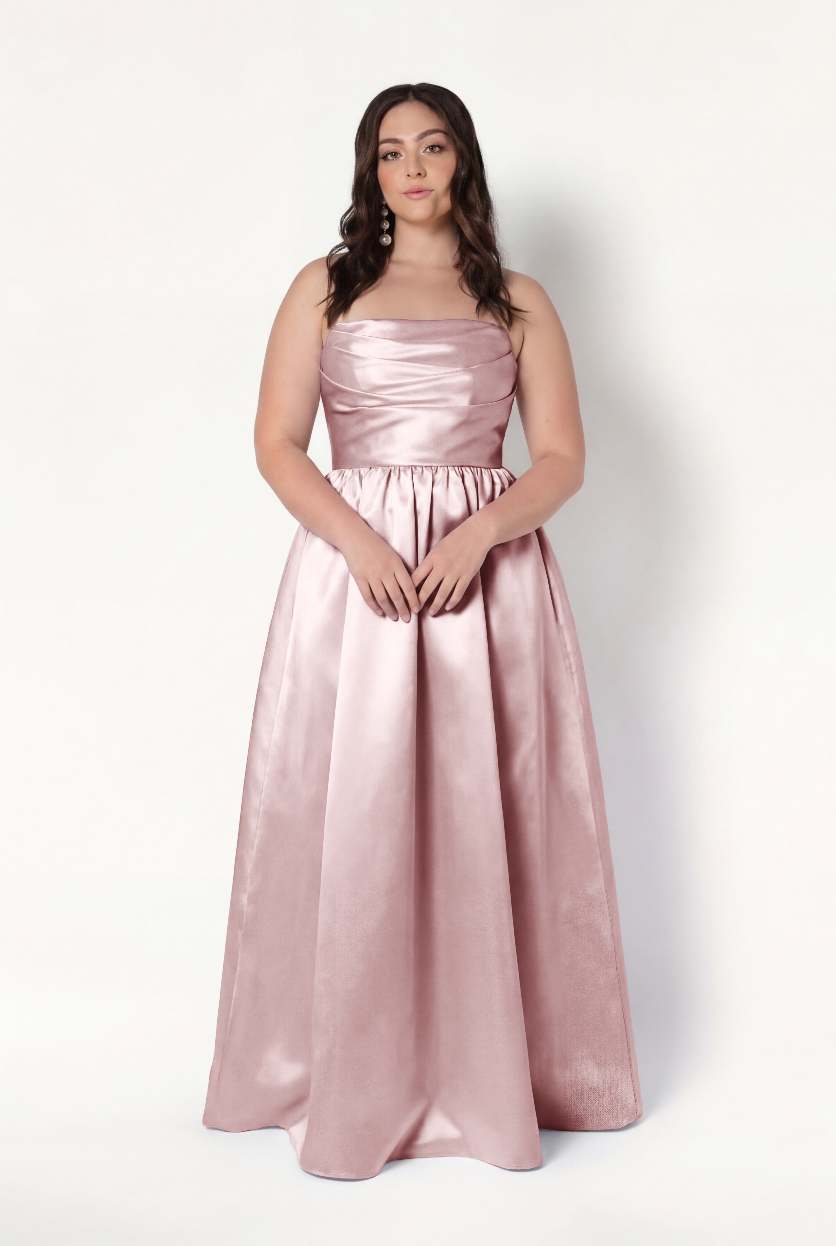 A woman with long, wavy brown hair wears the TH&TH Blair Bridesmaid Dress in Smoked Blush Satin Twill, a sleeveless, floor-length gown with optional detachable puff sleeves. She stands against a plain white background, hands on the skirt.