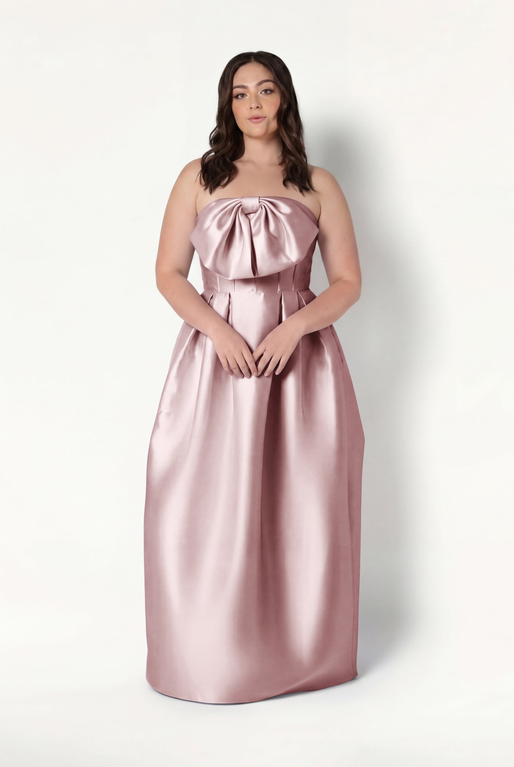 A woman with long brown hair wears the TH&TH Ophelia Bridesmaid Dress in smoked blush satin twill, featuring a strapless corset bodice and large bow, standing against a plain white background.