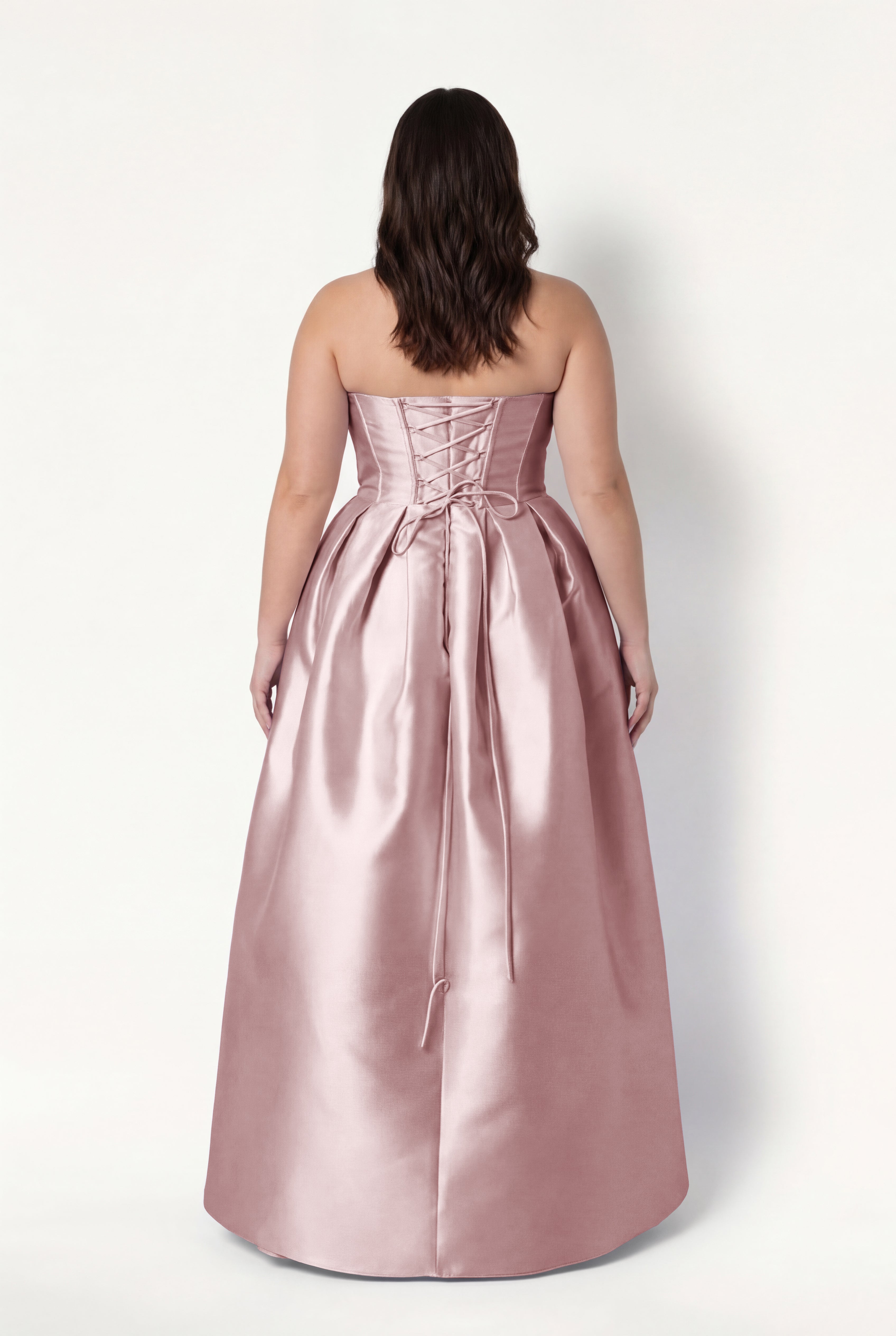 A woman with wavy brown hair wears the TH&TH Ophelia Bridesmaid Dress in Smoked Blush Satin Twill, featuring a strapless lace-up corset bodice, long ribbon ties, and floor-length skirt. She faces away against a plain white background.