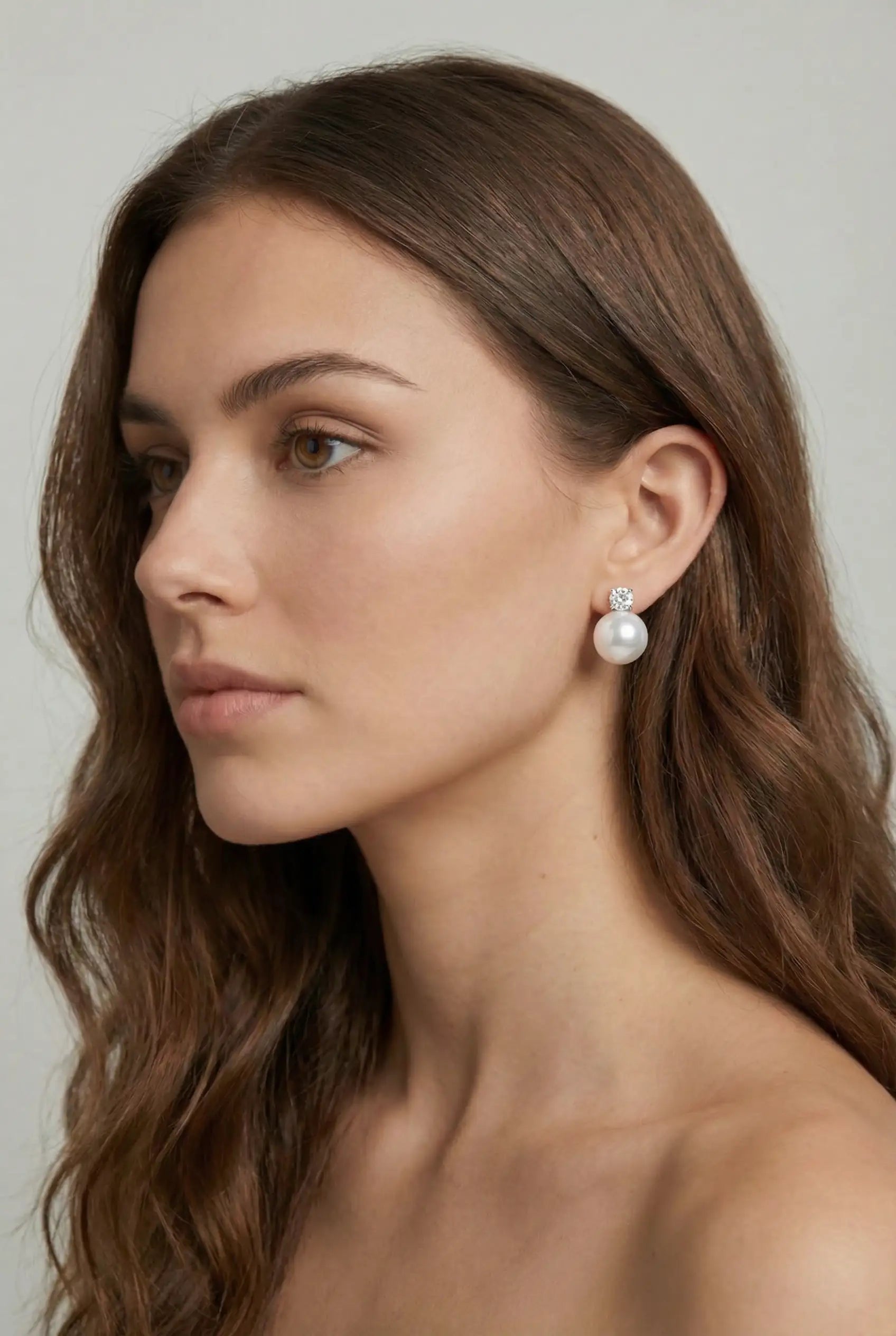 A woman with long, wavy brown hair and bare shoulders is shown in profile, wearing TH&TH's Solace Stud Earrings—elegant pearl crystal studs ideal for brides seeking handmade UK bridal jewellery—against a neutral background.