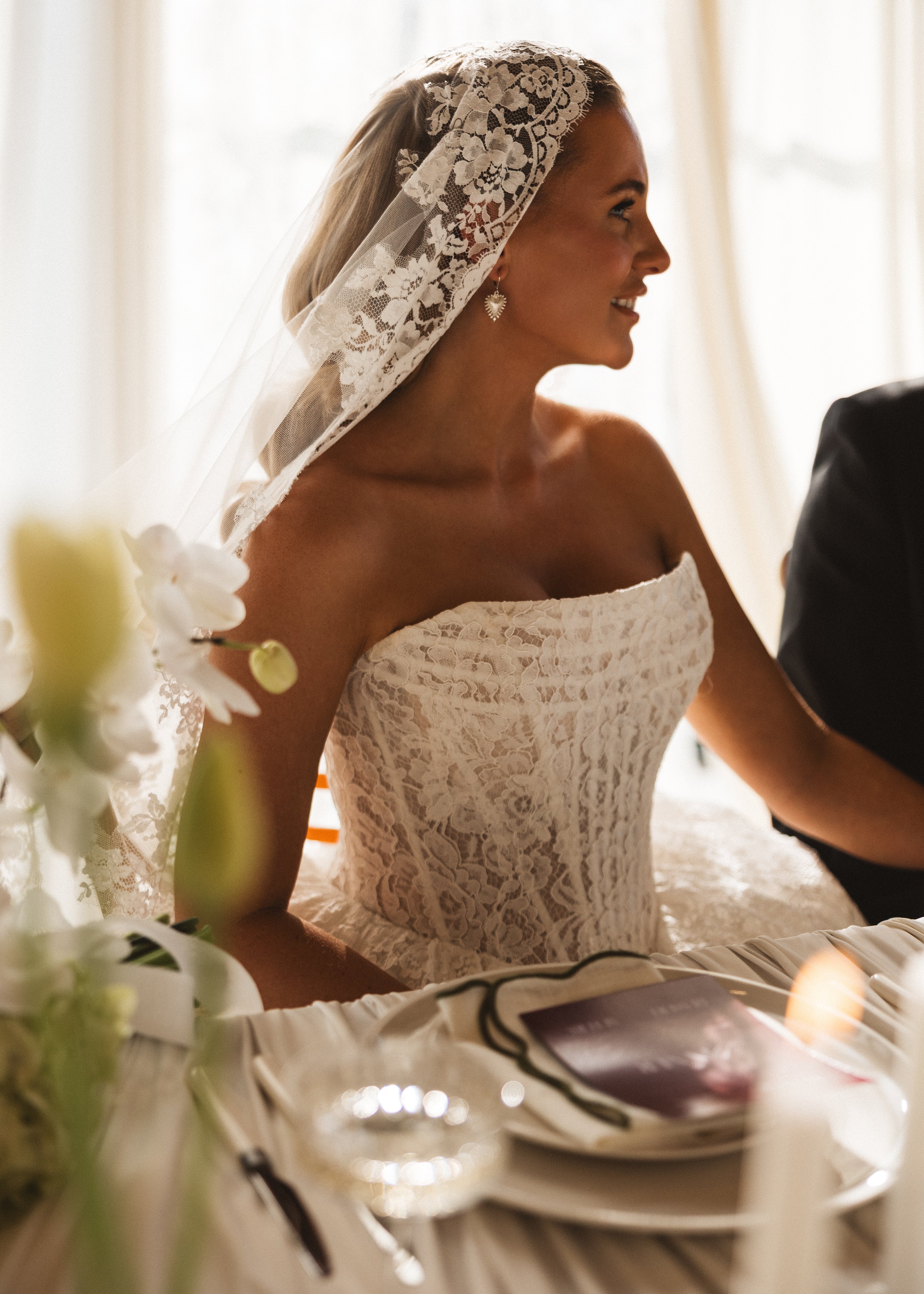 A bride in a lace wedding dress and the Catherine Eyelash Lace Mantilla Veil by TH&TH sits at a decorated table, smiling in soft natural light. White flowers and elegant table settings adorn the foreground.