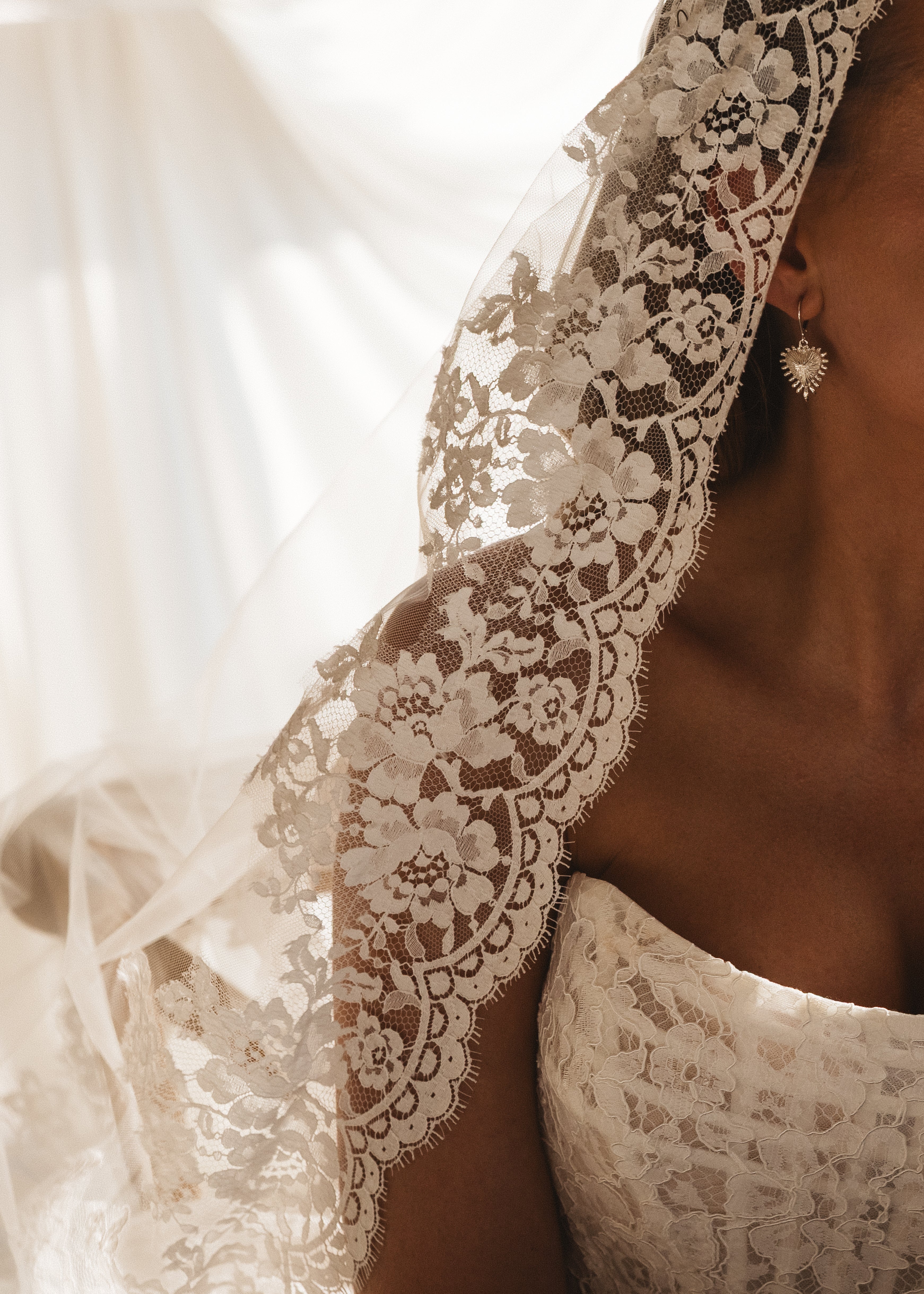 A bride wearing the TH&TH Catherine Eyelash Lace Mantilla Veil and heart-shaped earrings, in a strapless lace dress. Sunlight highlights the floral patterns as she turns her face away from the camera.