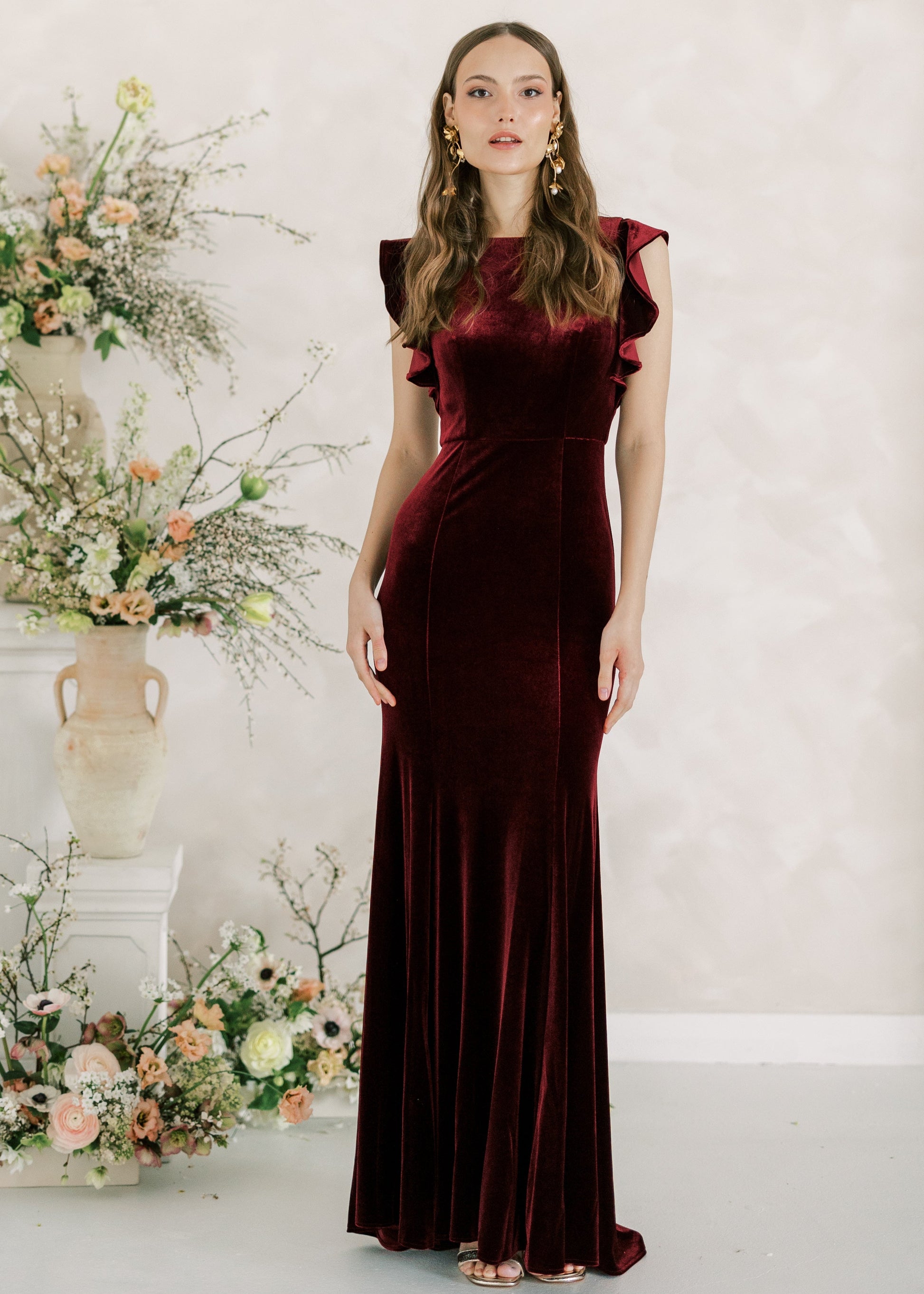 Woman in a burgundy evening gown standing in front of floral arrangements against a light background