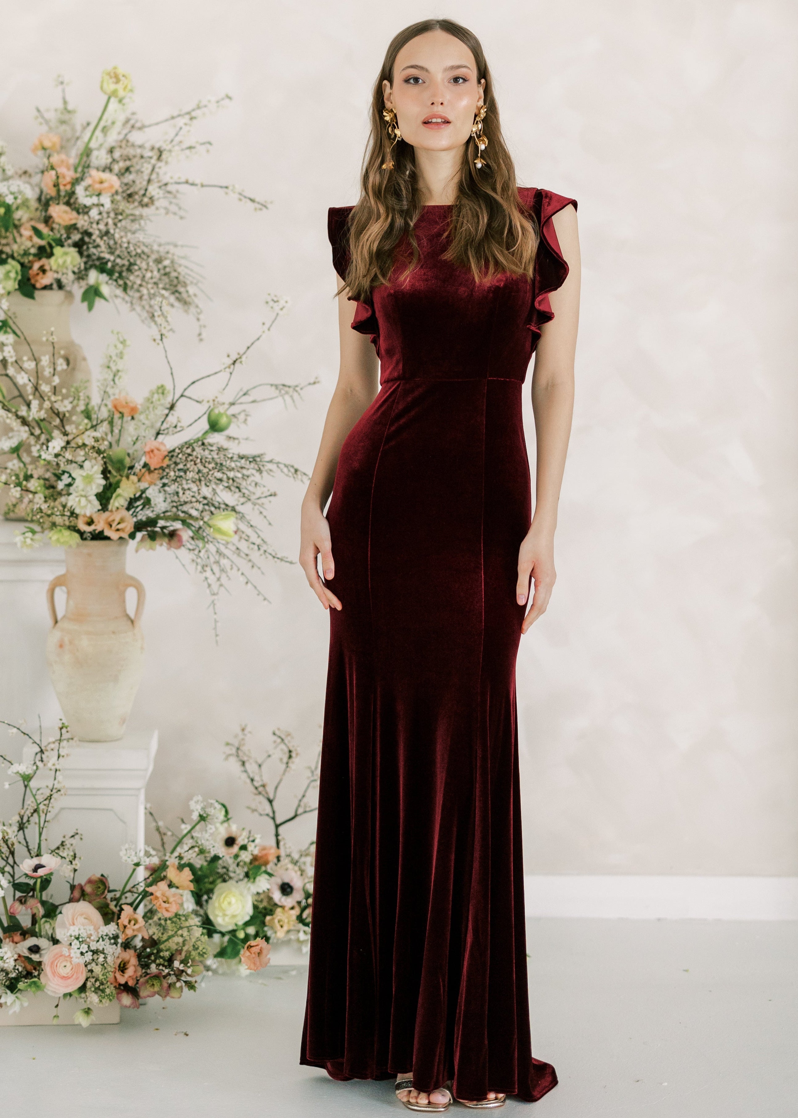 Woman in a burgundy evening gown standing in front of floral arrangements against a light background