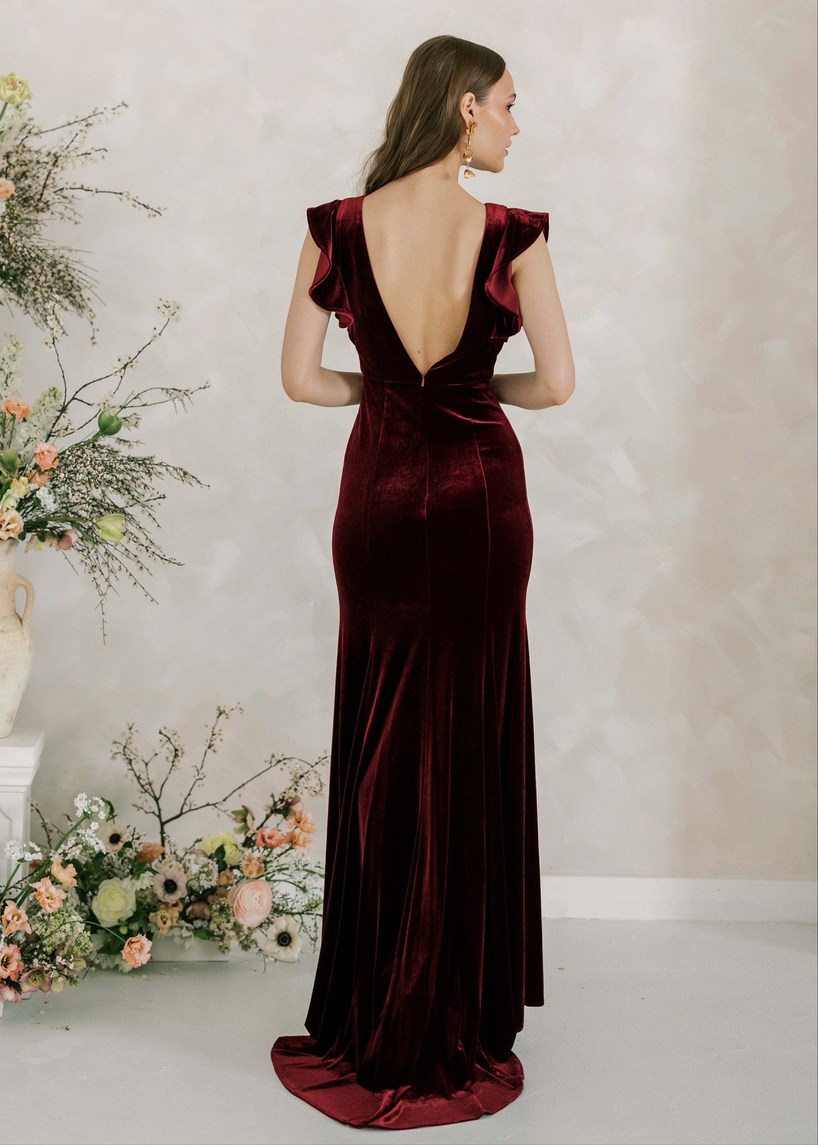 Woman in a burgundy evening gown standing in front of floral arrangements against a plain wall.