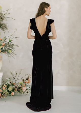 Woman in a black evening gown standing in front of floral arrangements against a plain wall.