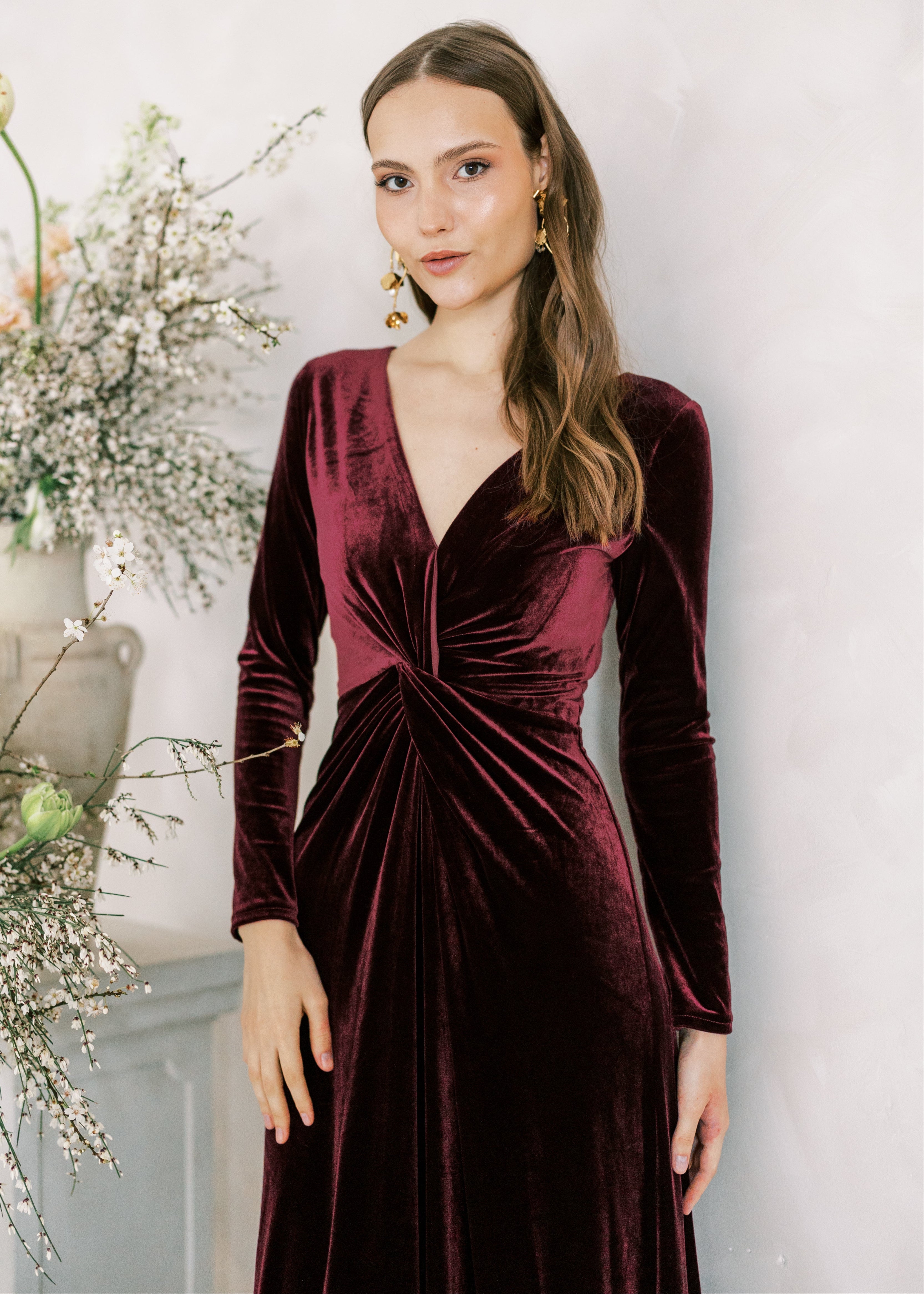 Woman wearing a burgundy velvet dress with long sleeves against a white wall.