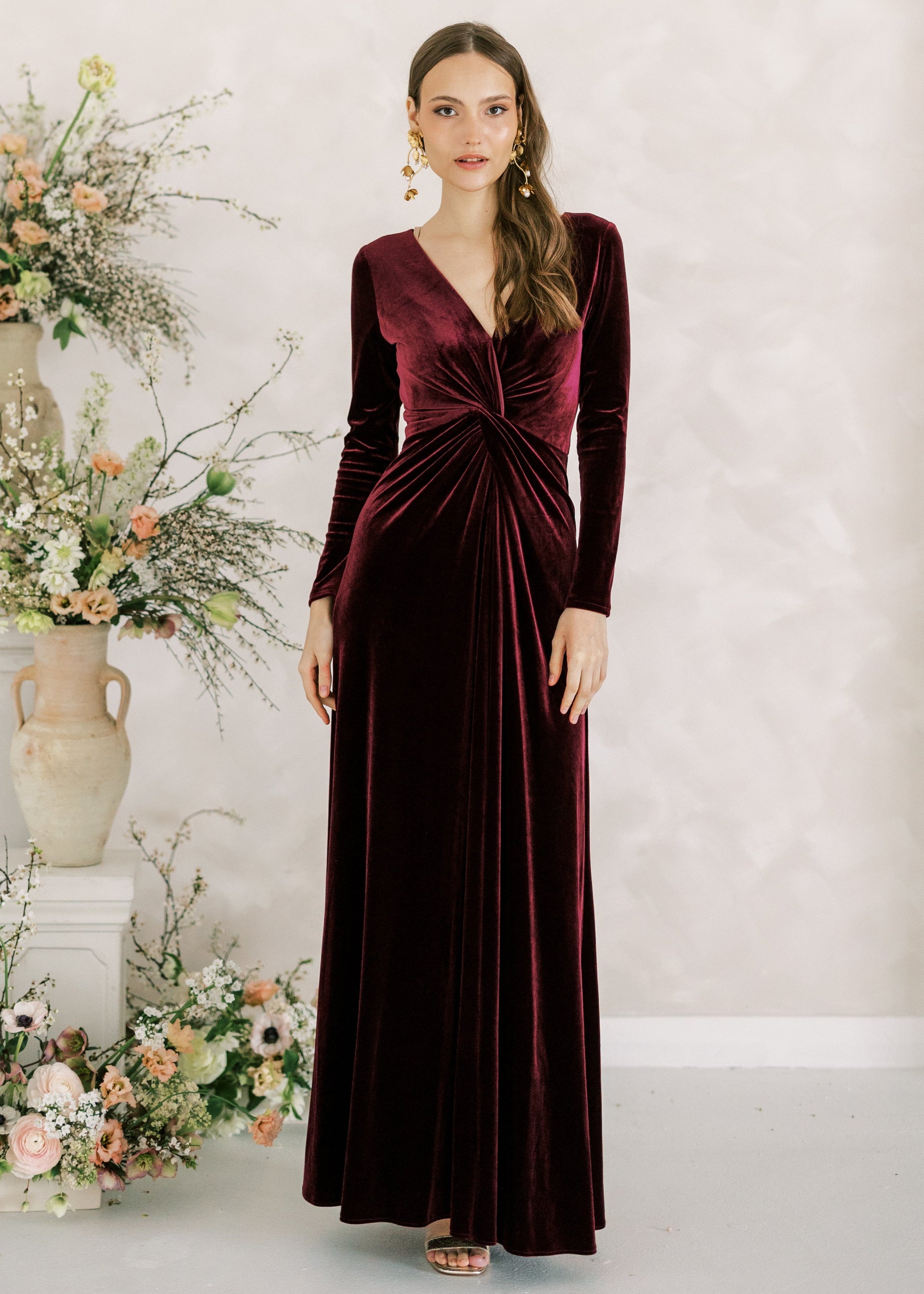 Woman in a burgundy velvet dress standing next to floral arrangements against a light background