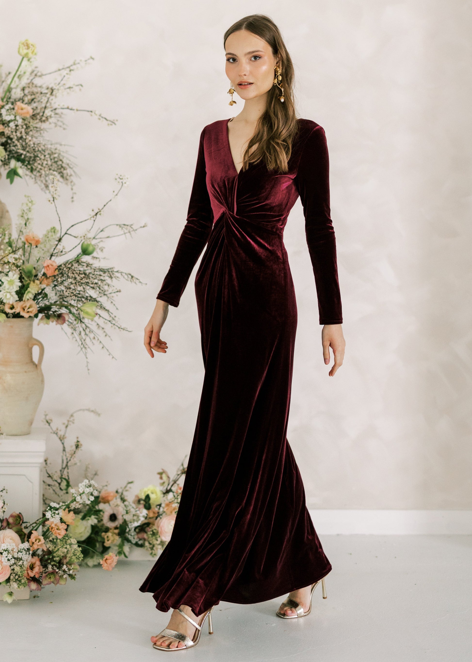 Woman in a burgundy velvet dress standing next to floral arrangements against a light background