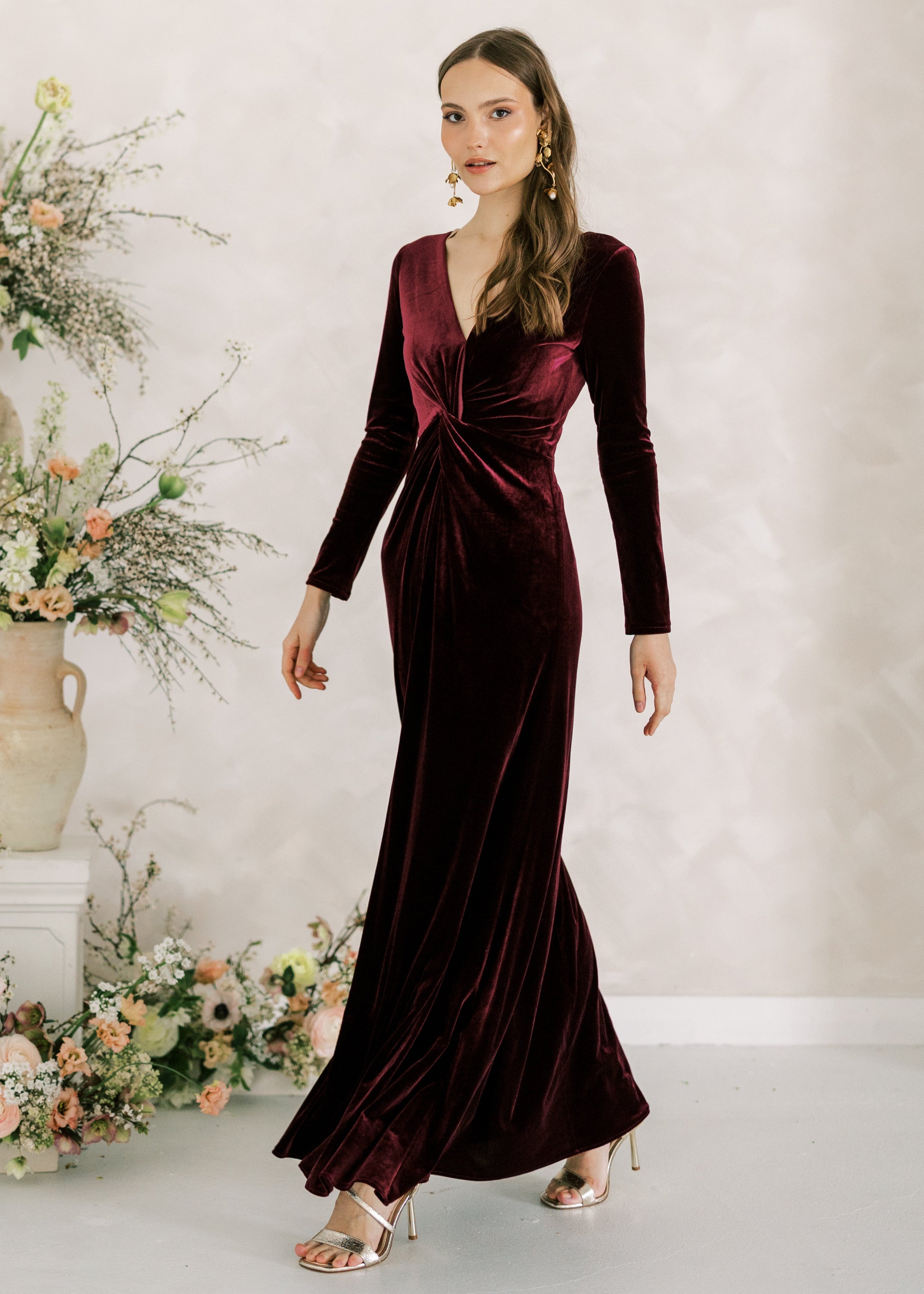 Woman in a burgundy velvet dress standing next to floral arrangements against a light background