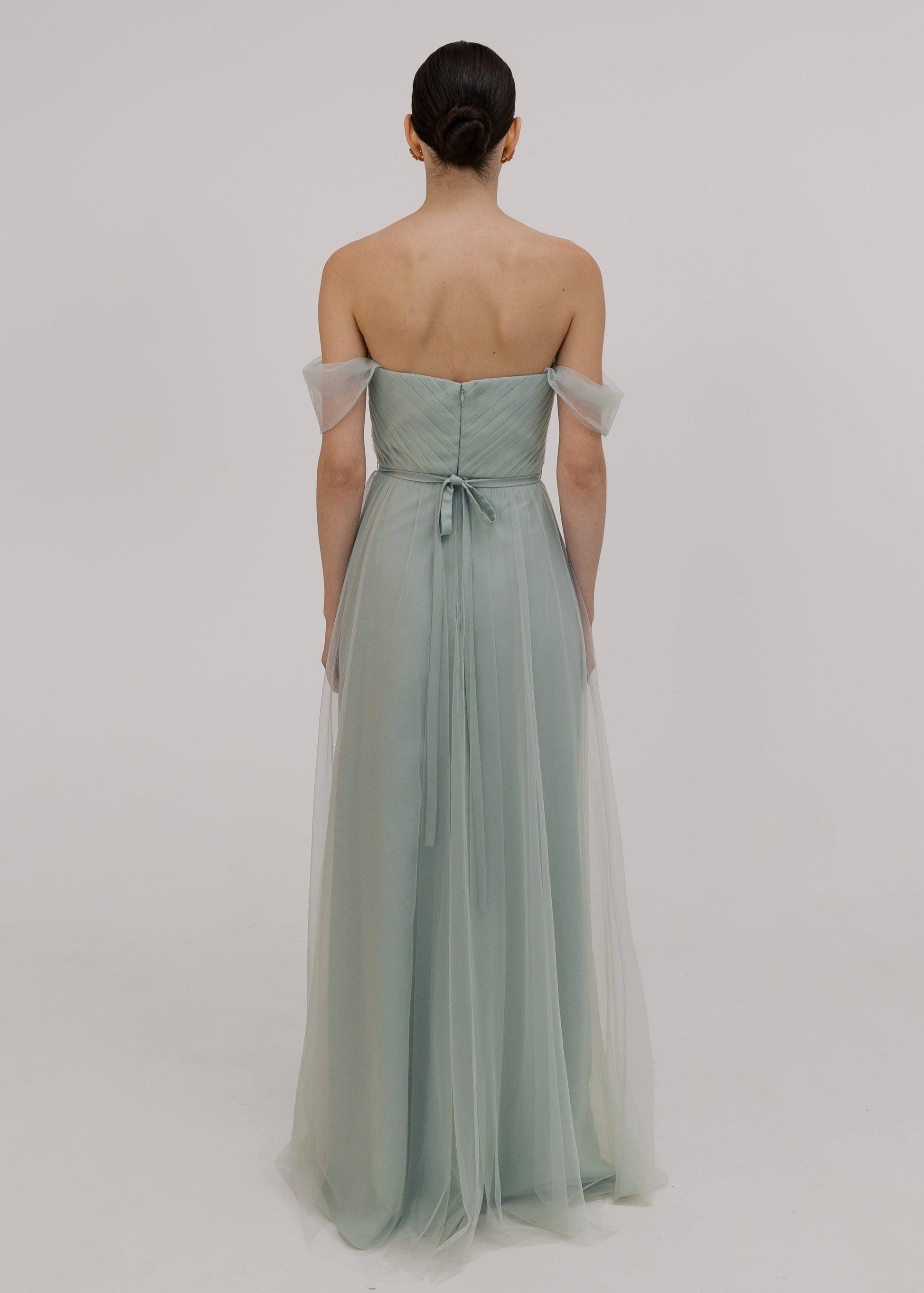 Woman wearing a light green strapless gown with sheer sleeves on a plain background back view with bow by TH&TH bridesmaids. 