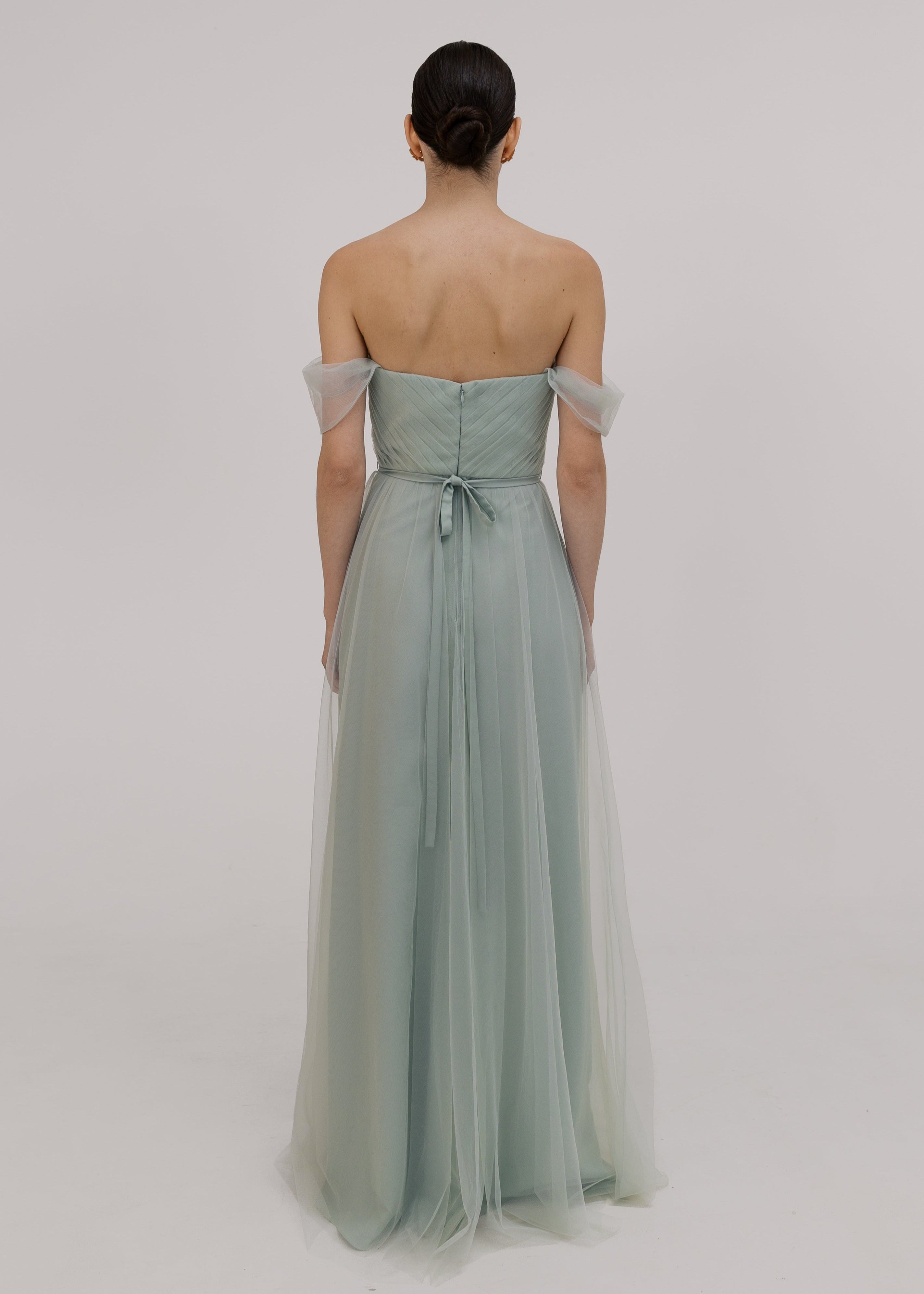 Woman wearing a light green strapless gown with sheer sleeves on a plain background back view with bow by TH&TH bridesmaids. 