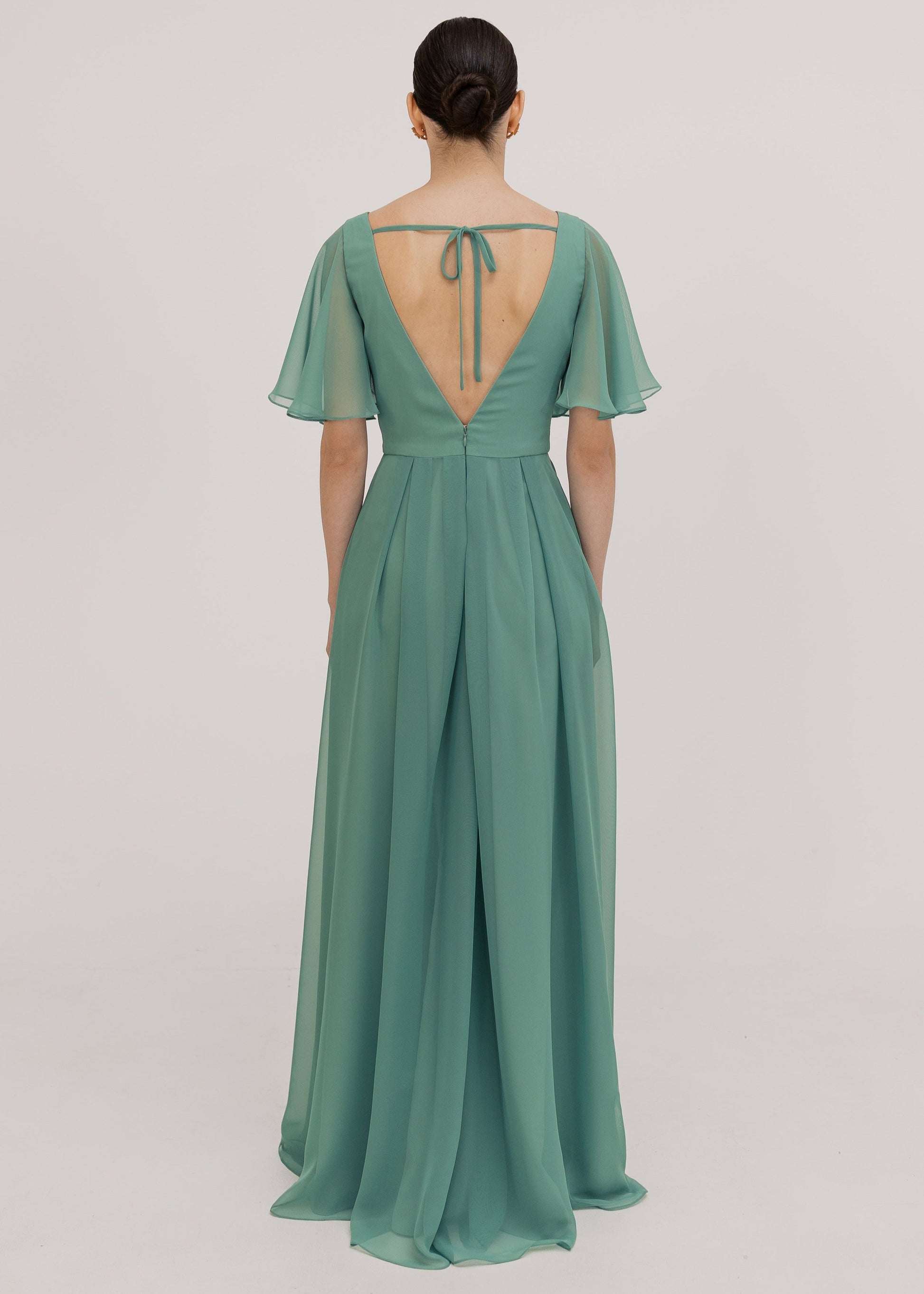 Model wears a sage green bridesmaid dress in chiffon with low v back details, flutter sleeves and flowing chiffon skirt by TH&TH Bridesmaids. 