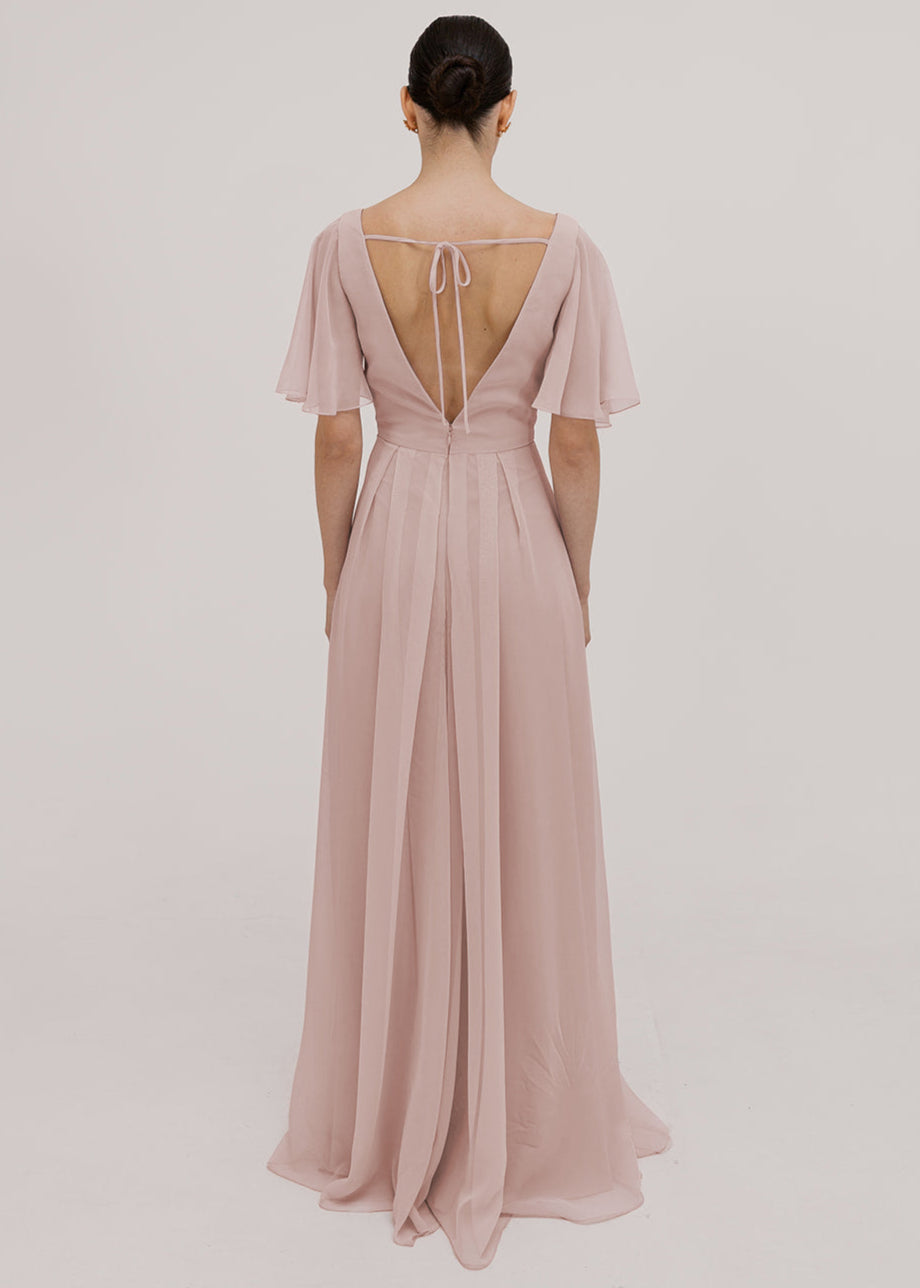Model wears a blush pink bridesmaid dress in chiffon with low back detail, flutter sleeves and flowing chiffon skirt by TH&TH Bridesmaids. 
