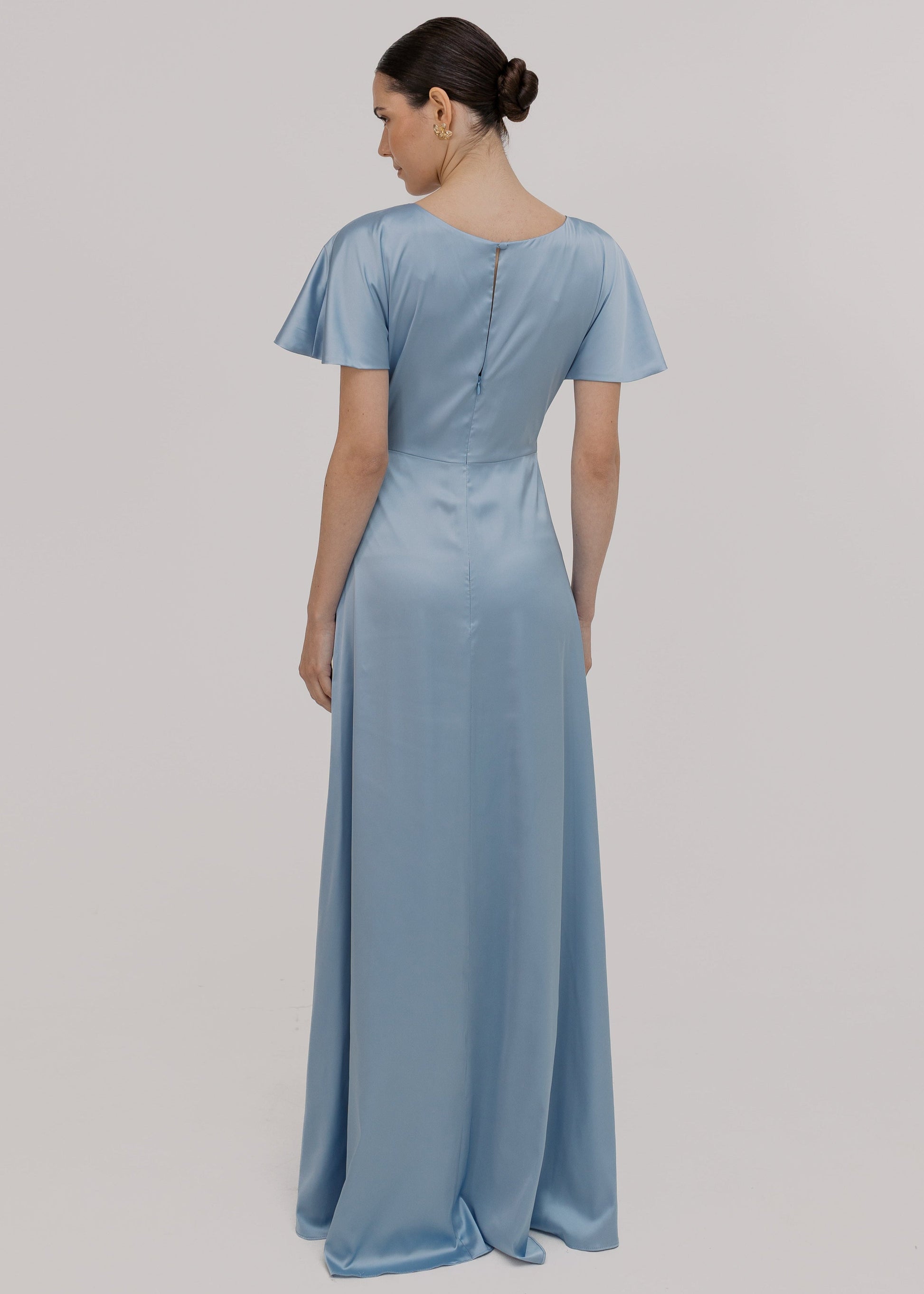 A woman wearing a dusty blue satin bridesmaid dress with v twist front neckline, flutter sleeves on a white background by TH&TH Bridesmaids.

