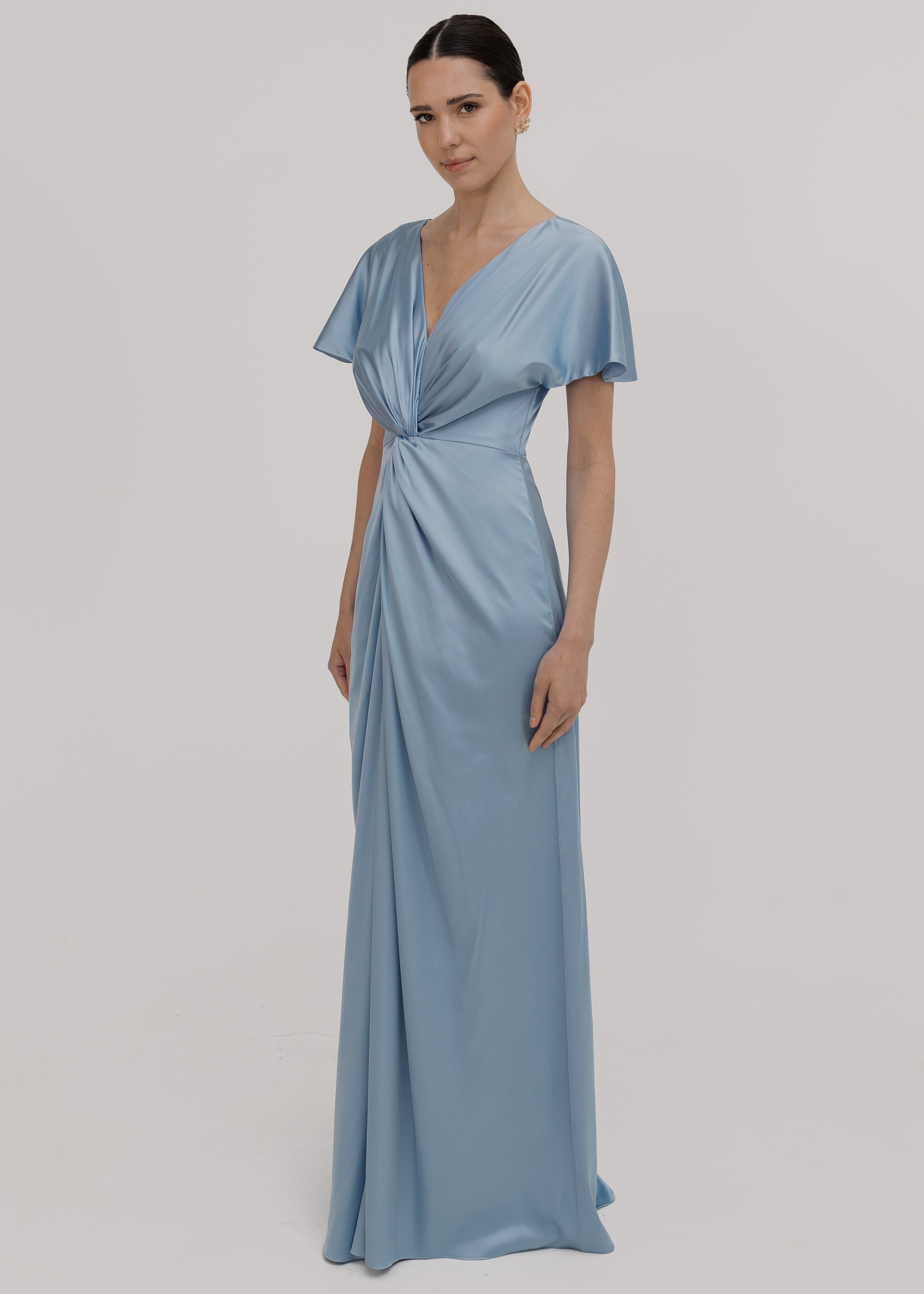 A woman wearing a dusty blue satin bridesmaid dress with v twist front neckline, flutter sleeves on a white background by TH&TH Bridesmaids.
