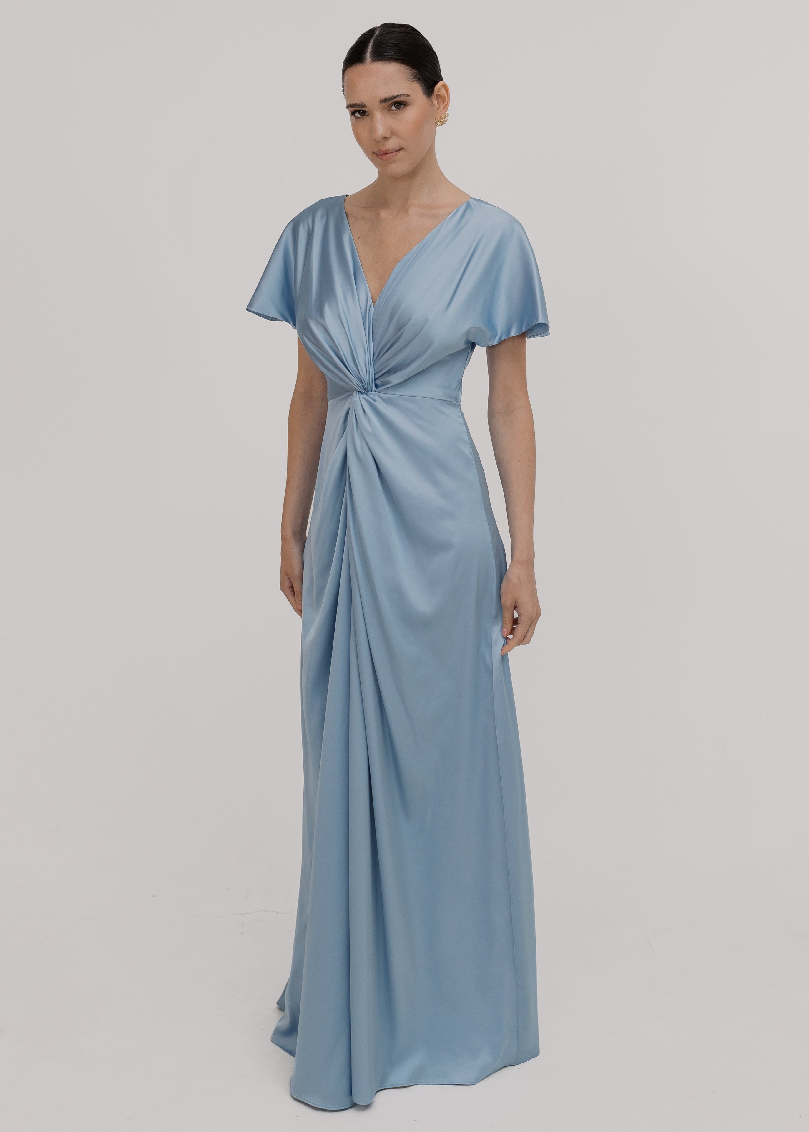 A woman wearing a dusty blue satin bridesmaid dress with v twist front neckline, flutter sleeves on a white background by TH&TH Bridesmaids.
