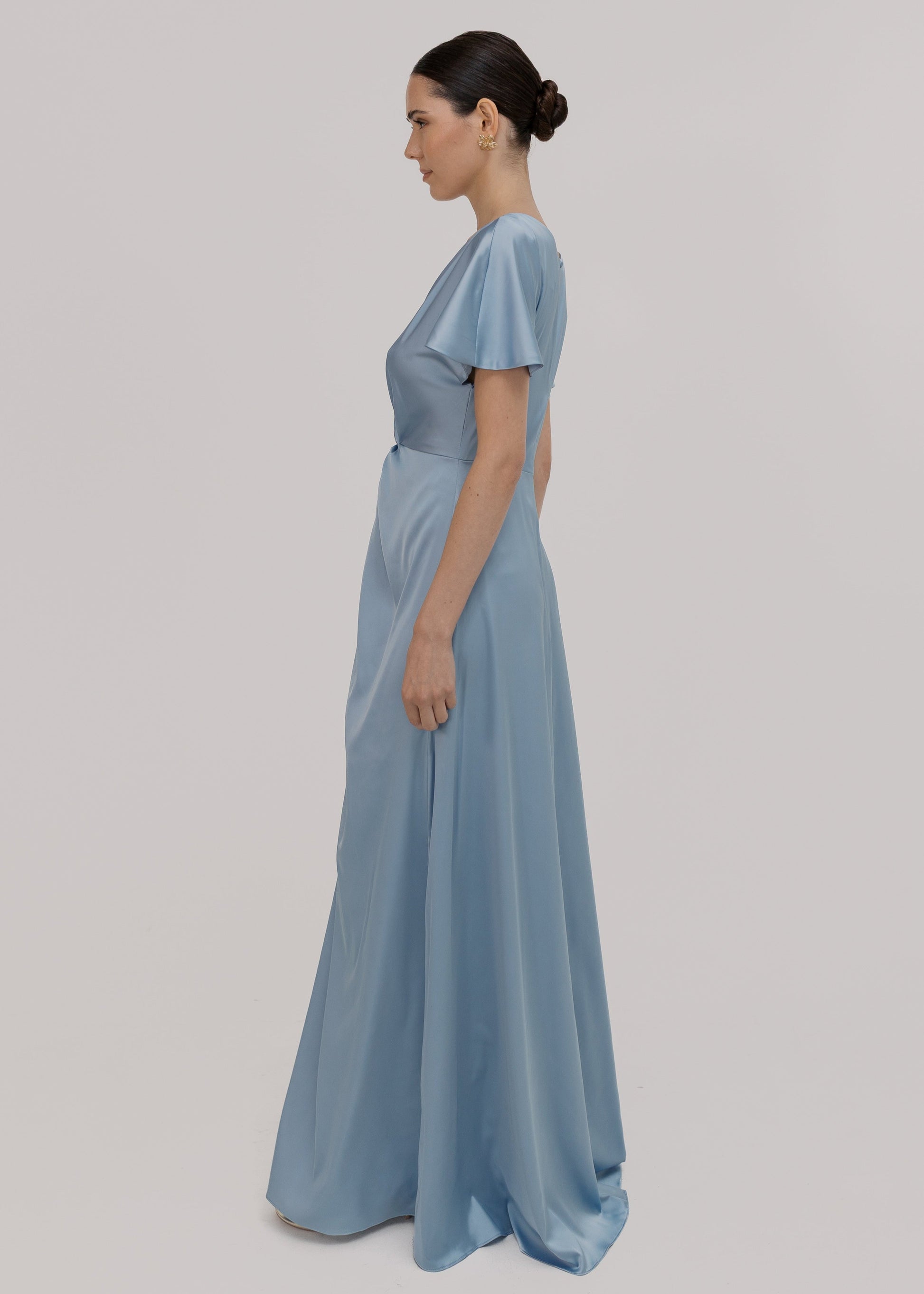 A woman wearing a dusty blue satin bridesmaid dress with v twist front neckline, flutter sleeves on a white background by TH&TH Bridesmaids.
