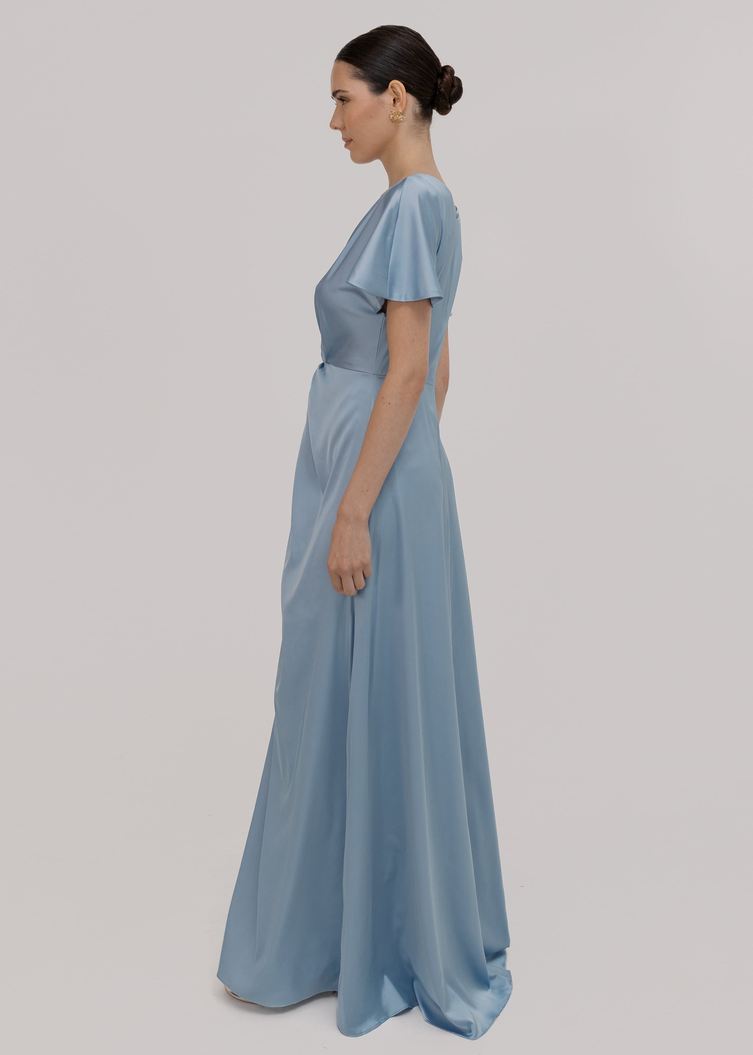 A woman wearing a dusty blue satin bridesmaid dress with v twist front neckline, flutter sleeves on a white background by TH&TH Bridesmaids.

