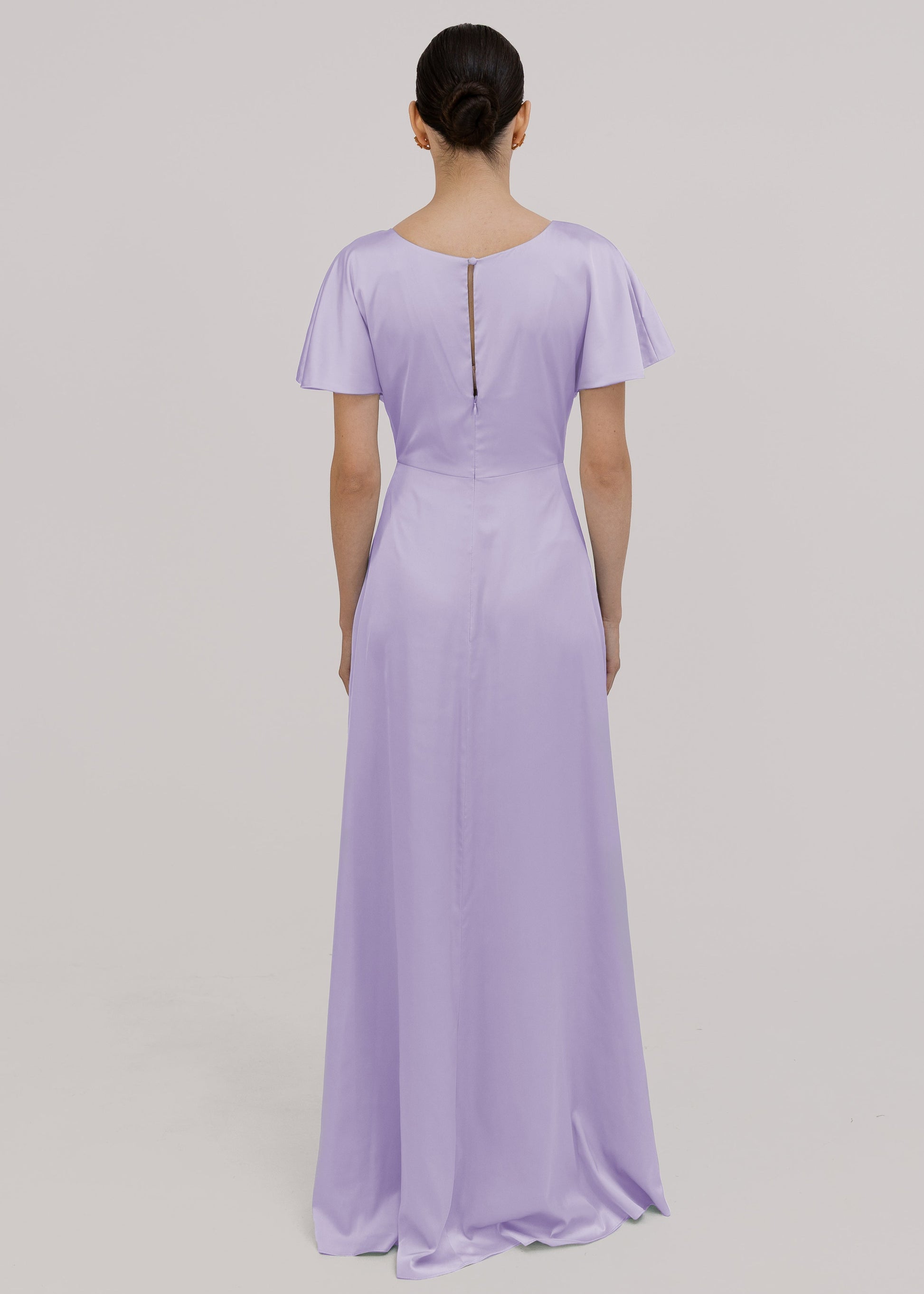 A woman wearing a lilac satin bridesmaid dress with flutter sleeves on a white background by TH&TH Bridesmaids. 
