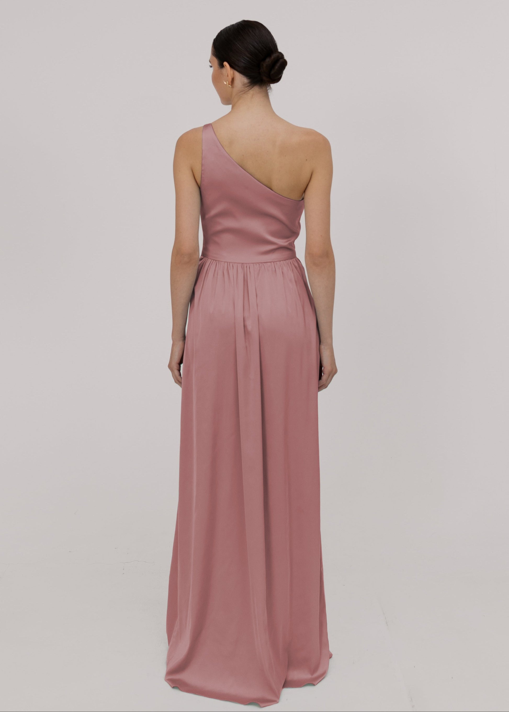 Model wearing a floor length pink satin bridesmaid dress with one shoulder and gathers in skirt by TH&TH Bridesmaids.

