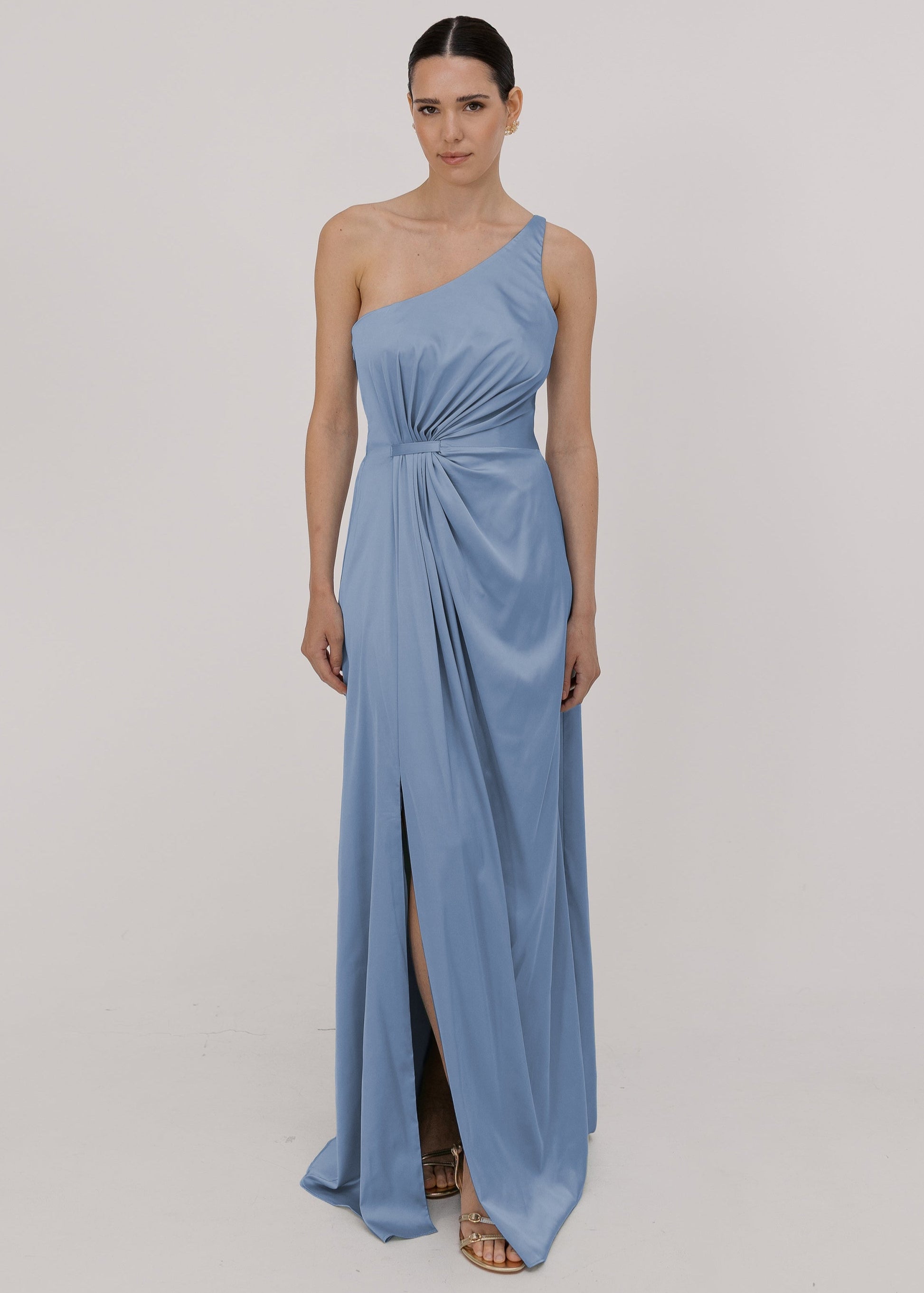Model wearing a floor length dusty blue satin bridesmaid dress with one shoulder and slit in skirt by TH&TH Bridesmaids.
