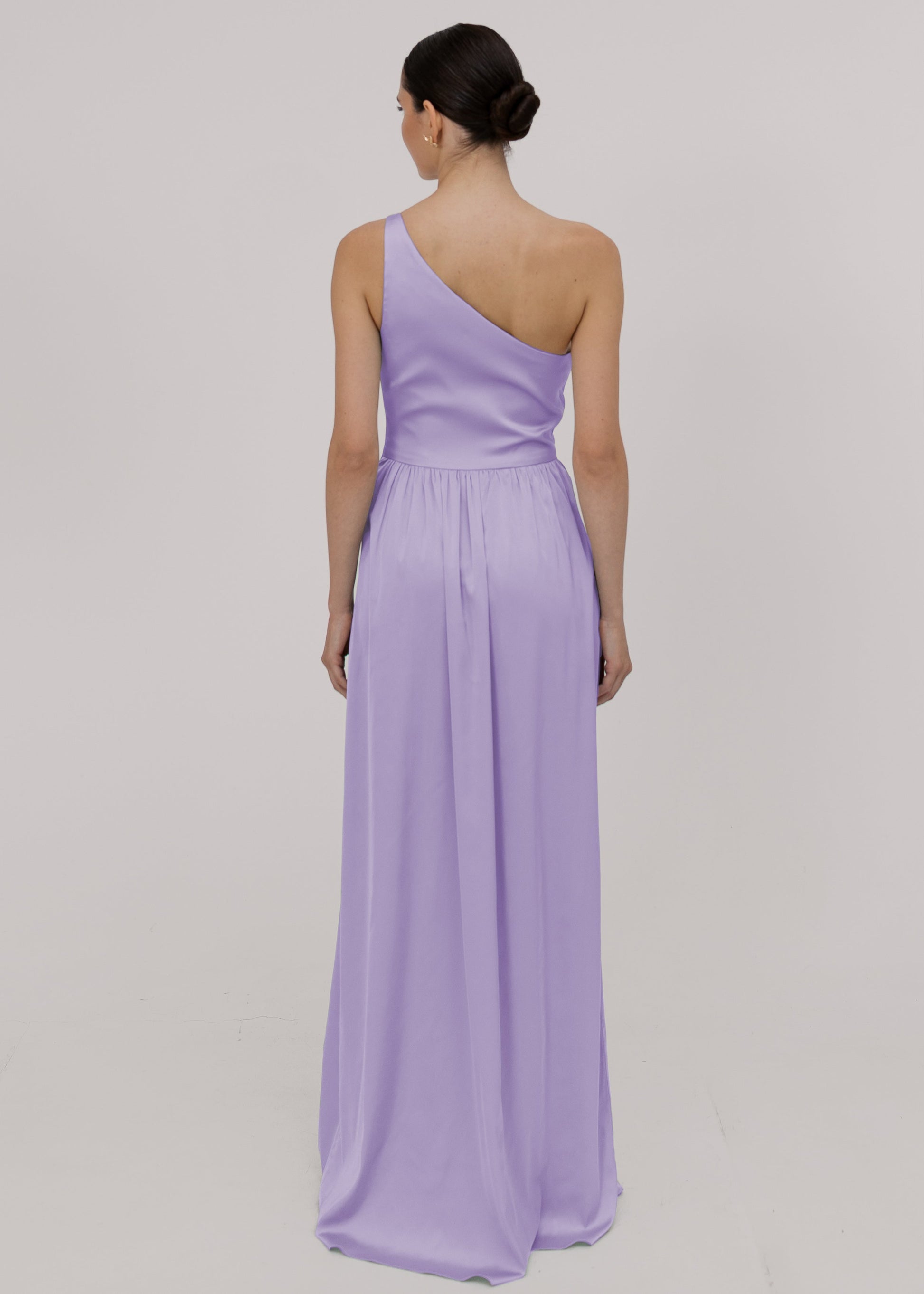 Model wearing a floor length lilac satin bridesmaid dress back view with one shoulder and slit in skirt by TH&TH Bridesmaids.
