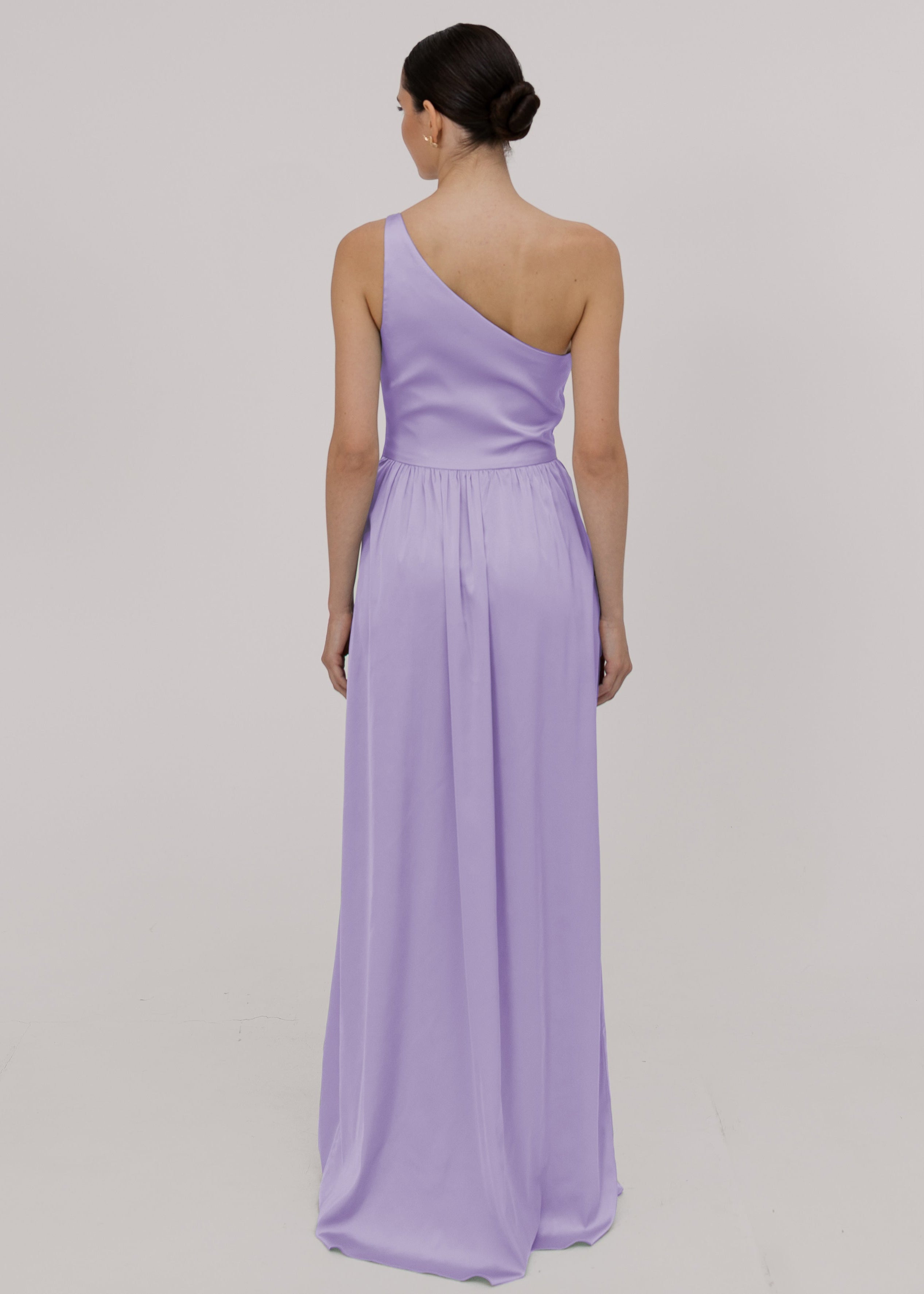 Model wearing a floor length lilac satin bridesmaid dress back view with one shoulder and slit in skirt by TH&TH Bridesmaids.
