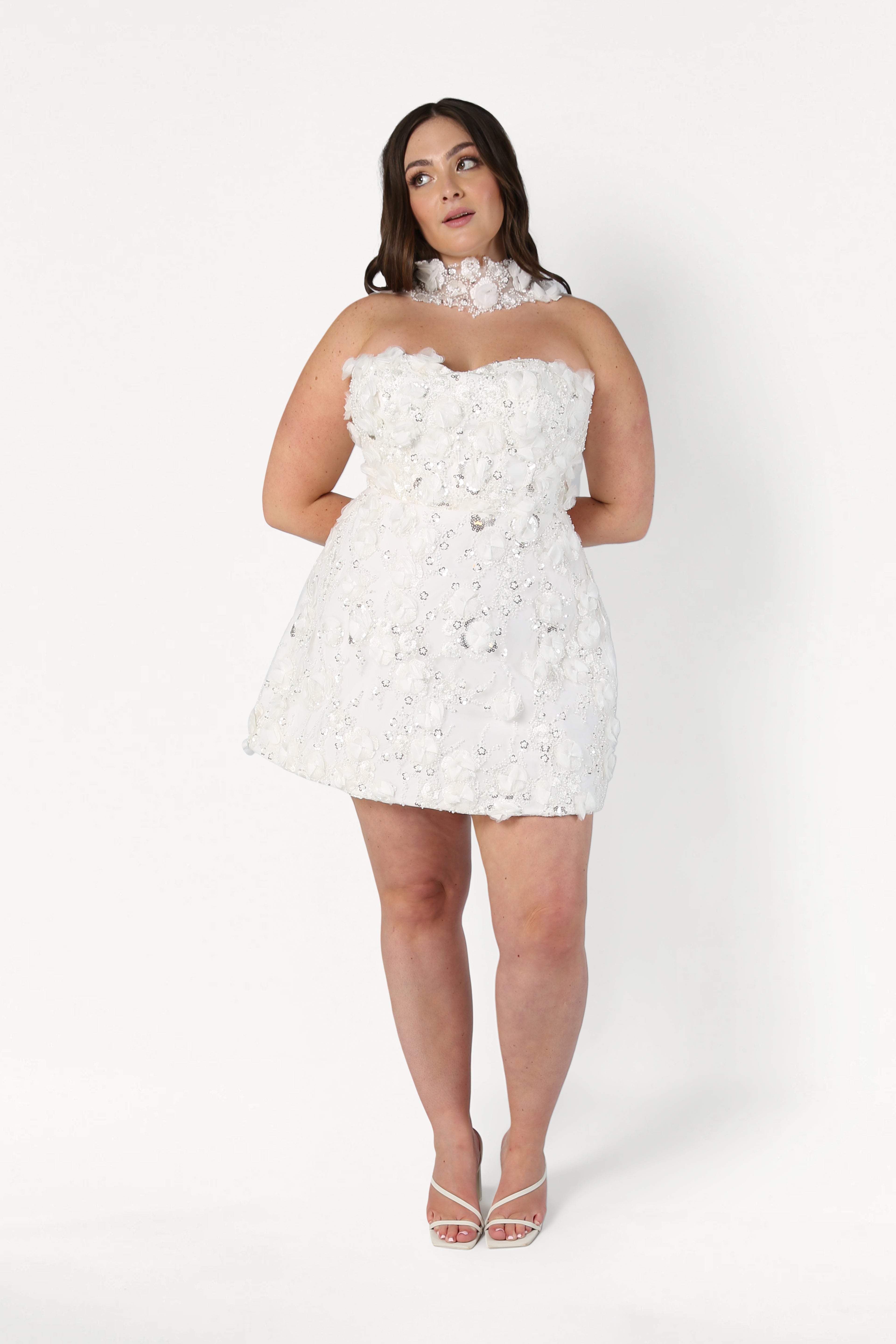 A woman models the TH&TH Taylor Bridal Mini Dress in Ivory, a short strapless design with 3D embellished tulle flowers and a matching floral choker, standing against a plain white background in white heels with hands behind her back.
