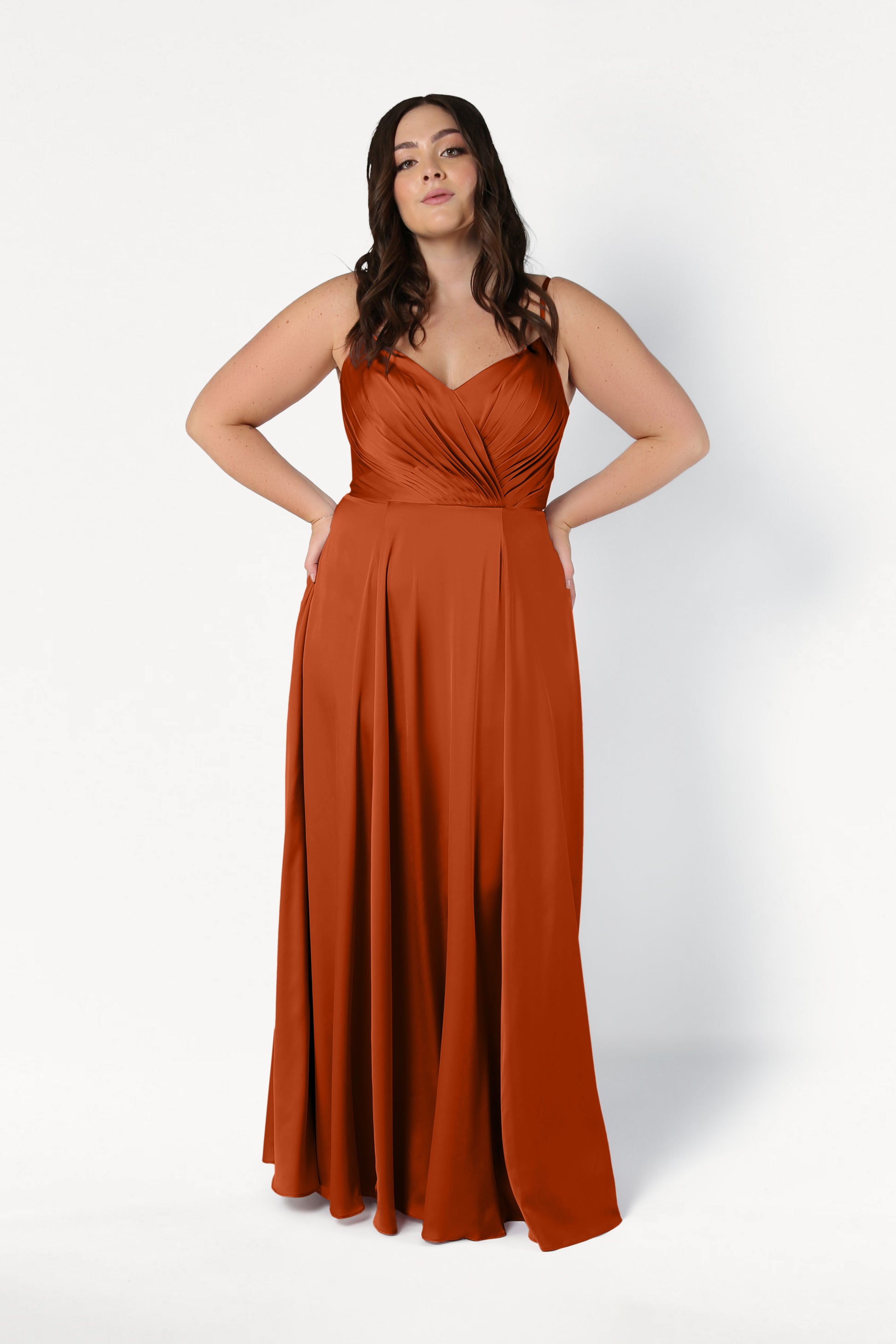 A woman with long brown hair wears the TH&TH Isla Bridesmaid Dress in Terracotta Satin, featuring a sleeveless, floor-length design with a fitted, pleated bodice and full skirt. She poses against a plain white background with her hands on her hips.