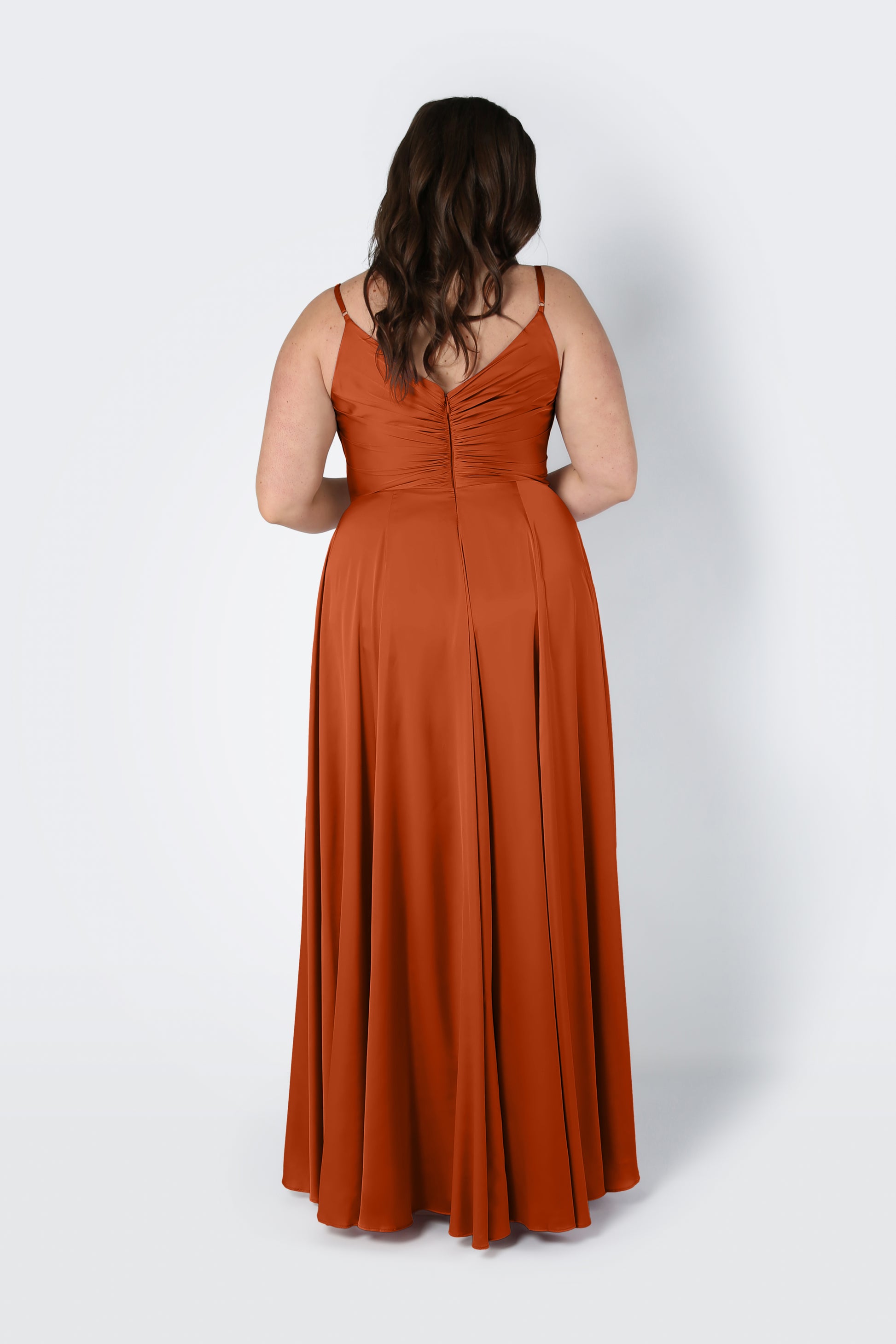 A woman with long brown hair models the TH&TH Isla Bridesmaid Dress in Terracotta Satin, a floor-length gown with thin straps and gathered detailing on the back, standing against a plain light background.