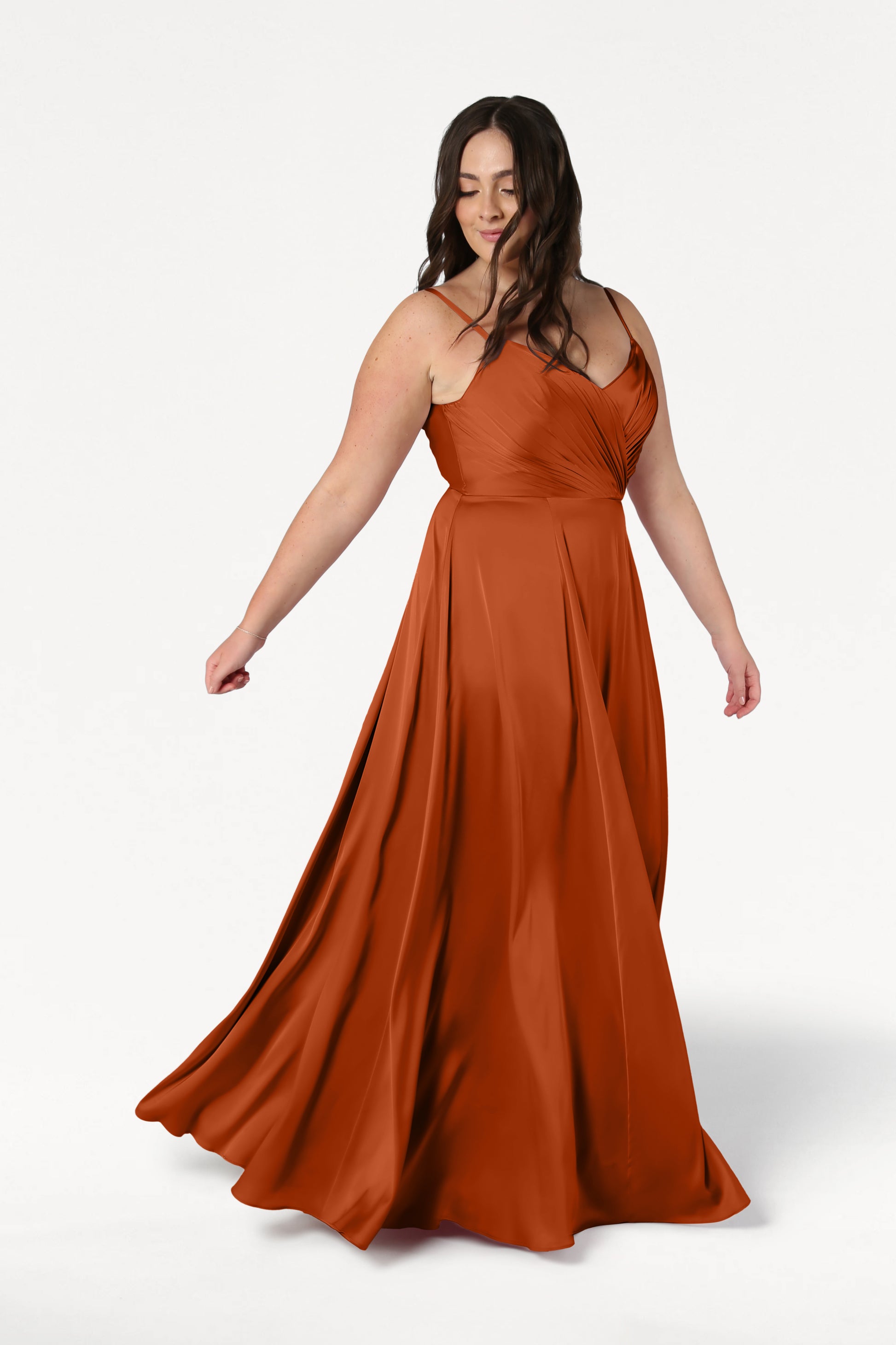 A woman with long brown hair gently twirls, creating movement in the TH&TH Isla Bridesmaid Dress in Terracotta Satin, which features a flowing silhouette and pleated bodice, set against a plain white background.