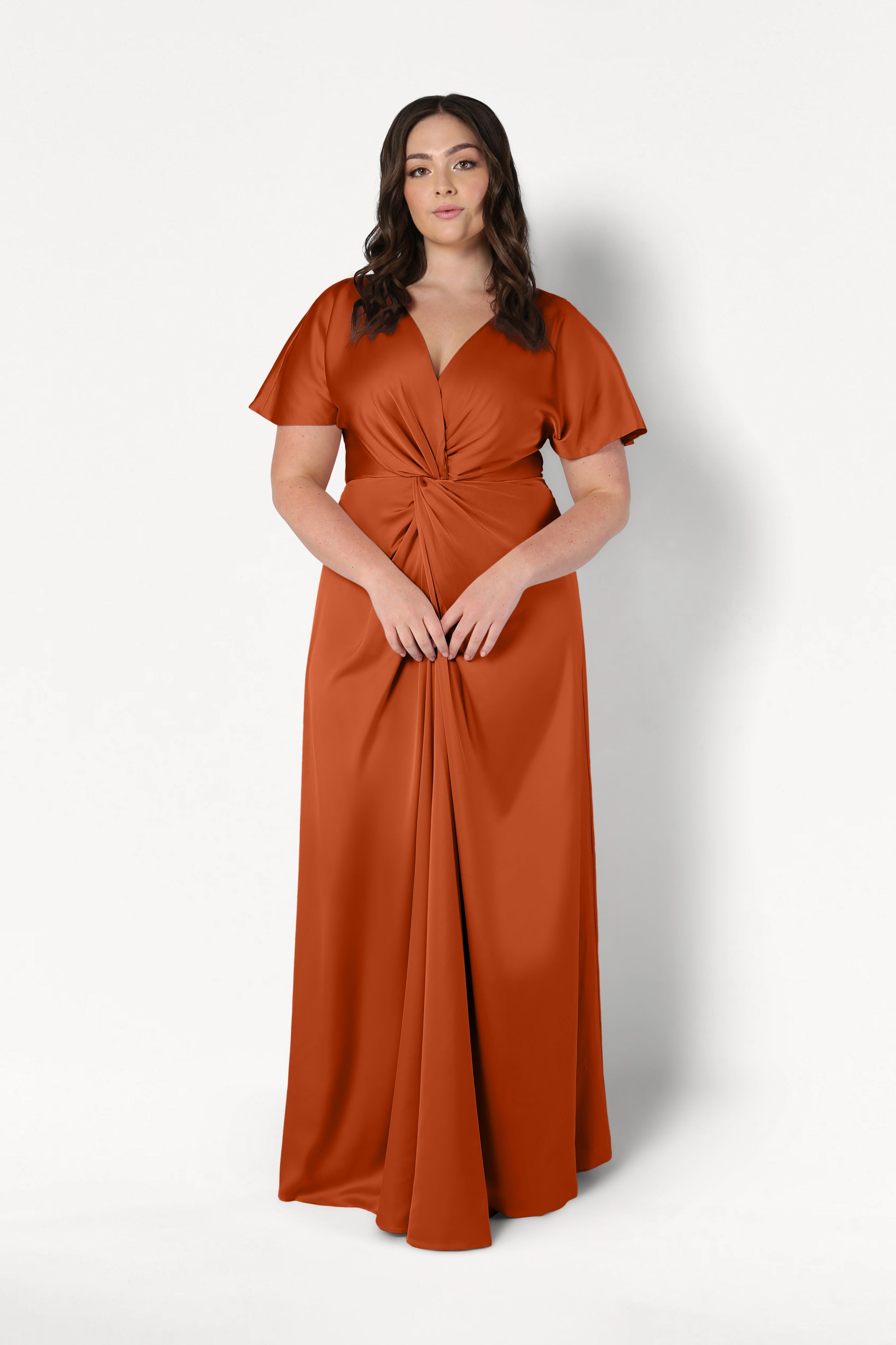 A woman with wavy brown hair wears the TH&TH Camilla Bridesmaid Dress in Terracotta Satin, a floor-length gown with a twist-knot bodice and short sleeves, standing against a plain white background.