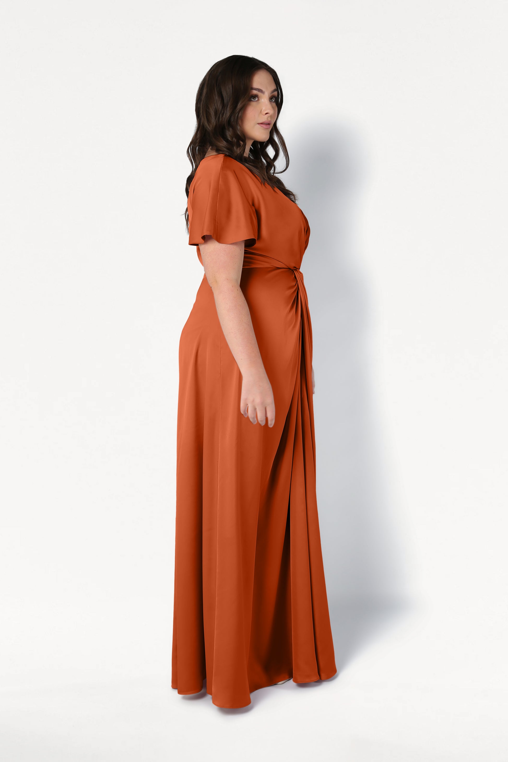 A woman with long dark hair stands in profile against a white background, wearing the size-inclusive TH&TH Camilla Bridesmaid Dress in Terracotta Satin, featuring short sleeves and a flowing silhouette.