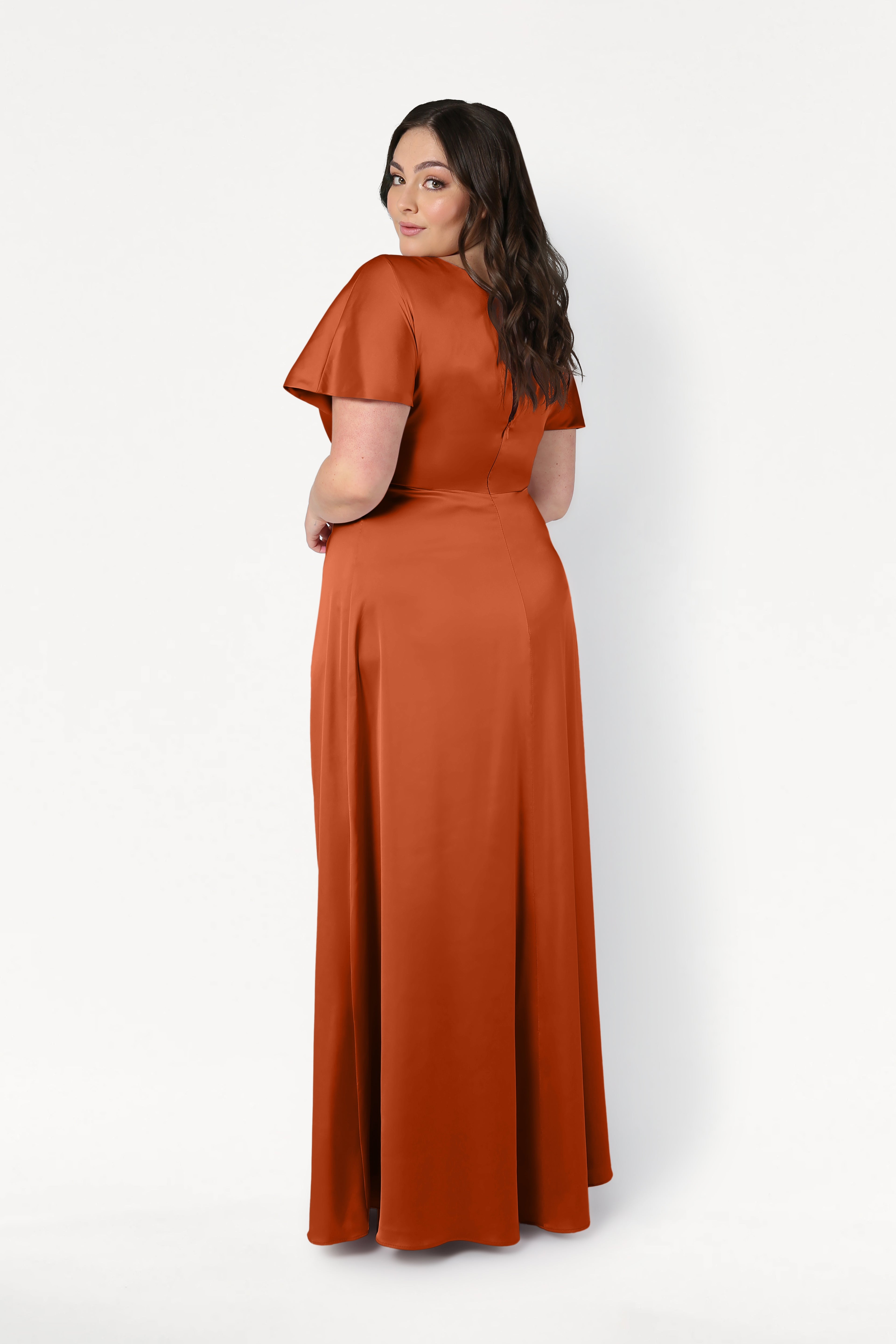 A woman with long, wavy dark hair faces away, looking over her shoulder. She wears the size-inclusive TH&TH Camilla Bridesmaid Dress in Terracotta Satin, a floor-length gown with short sleeves, against a plain white background.