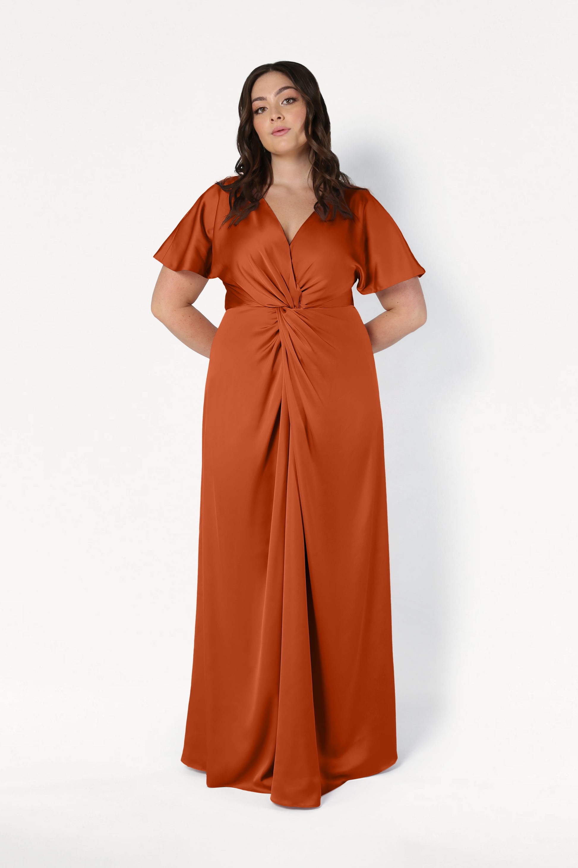 A woman with wavy brown hair models the TH&TH Camilla Bridesmaid Dress in Terracotta Satin—a size-inclusive, floor-length gown with short sleeves, a V-neckline, and gathered waist—against a plain white background.