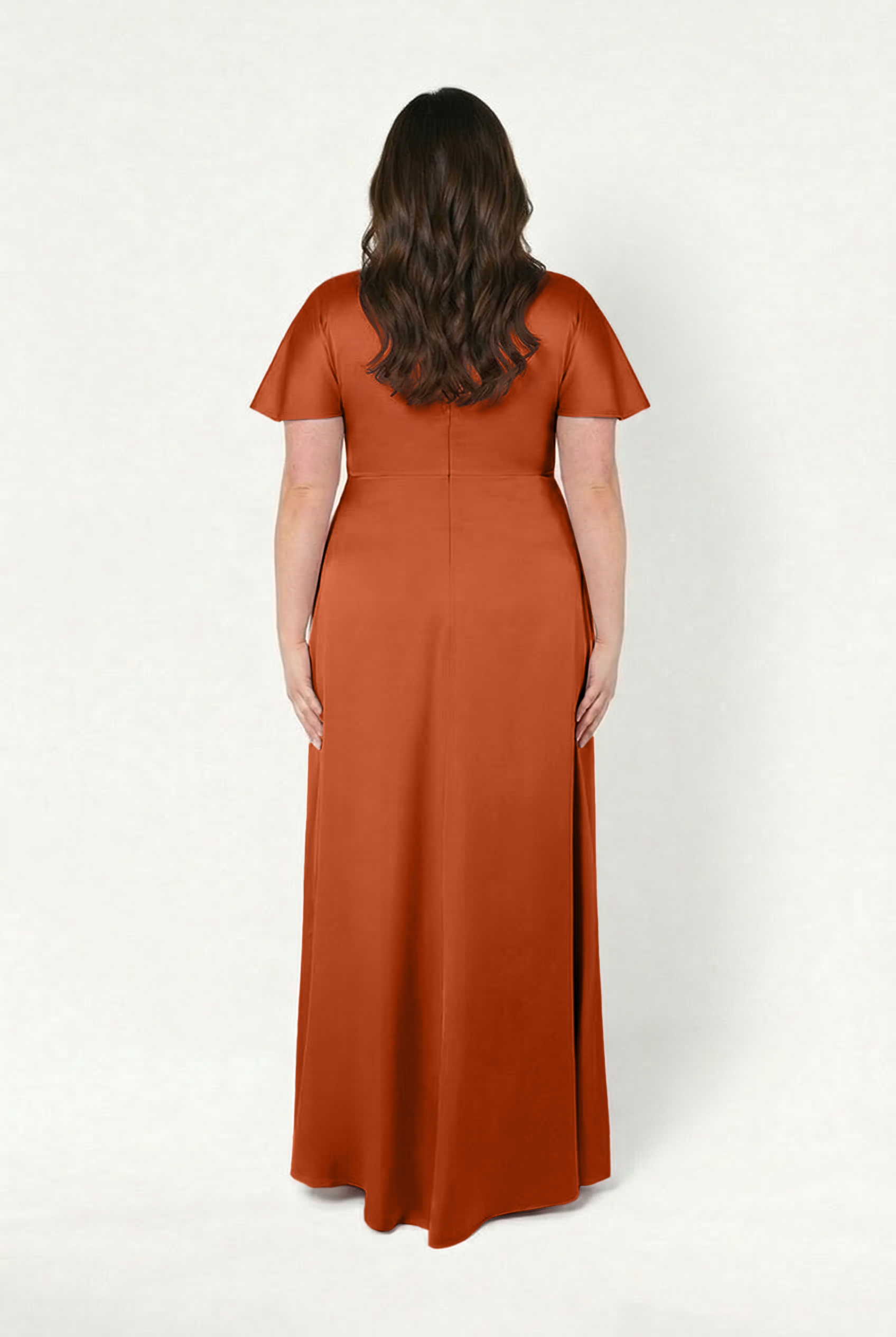 A woman with long brown hair wears the TH&TH Camilla Bridesmaid Dress in Terracotta Satin, a size-inclusive, floor-length dress with short sleeves, standing facing away against a plain white background.
