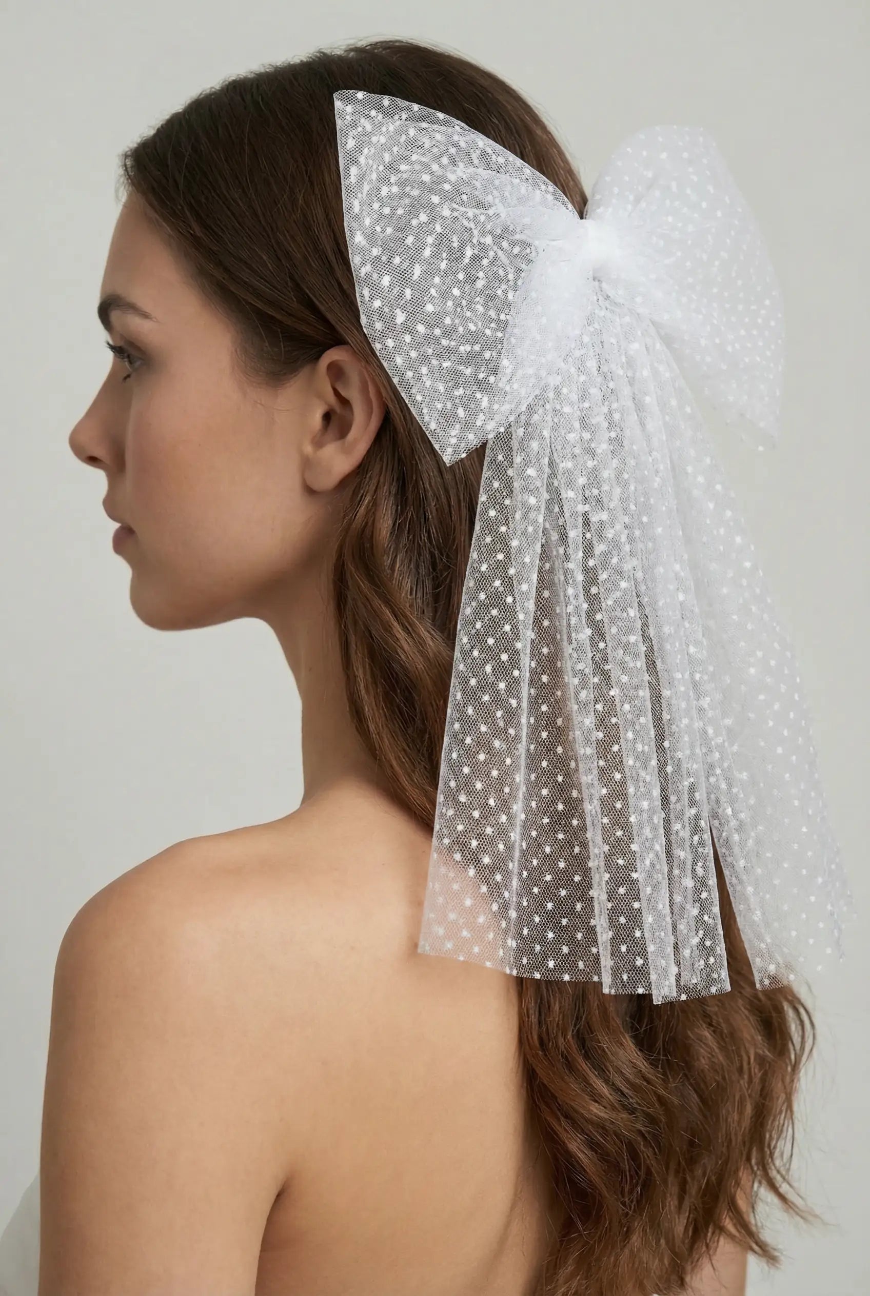 A woman with long brown hair, seen from the side, wears the TH&TH Thea Spot Tulle Bow Veil—a large white veil with trailing ribbons—adding whimsy to her bare shoulders against a plain light background.