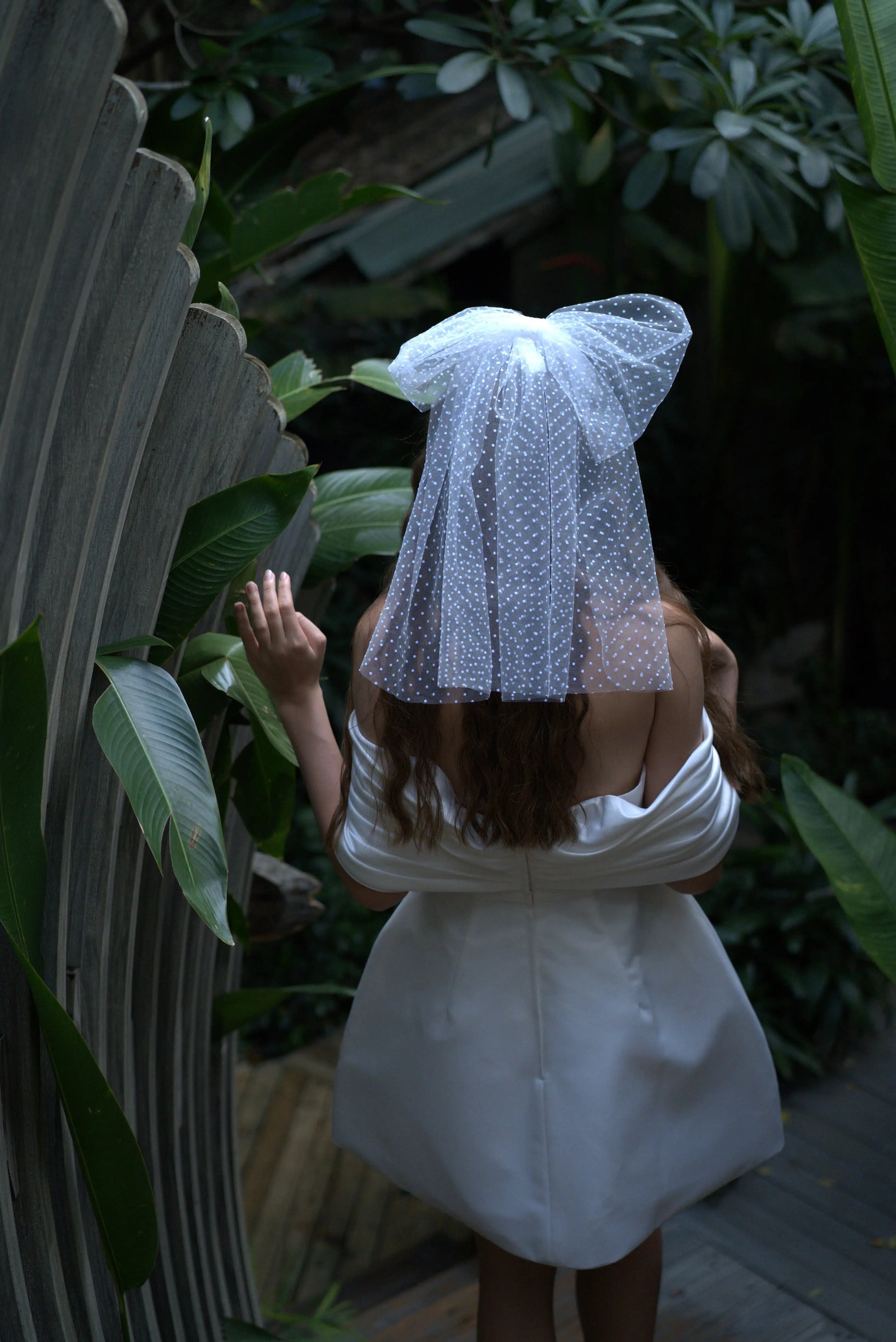 A woman in a short white off-shoulder dress wears the TH&TH Thea Spot Tulle Bow Veil, standing with her back to the camera among lush green plants and curved wooden structures.