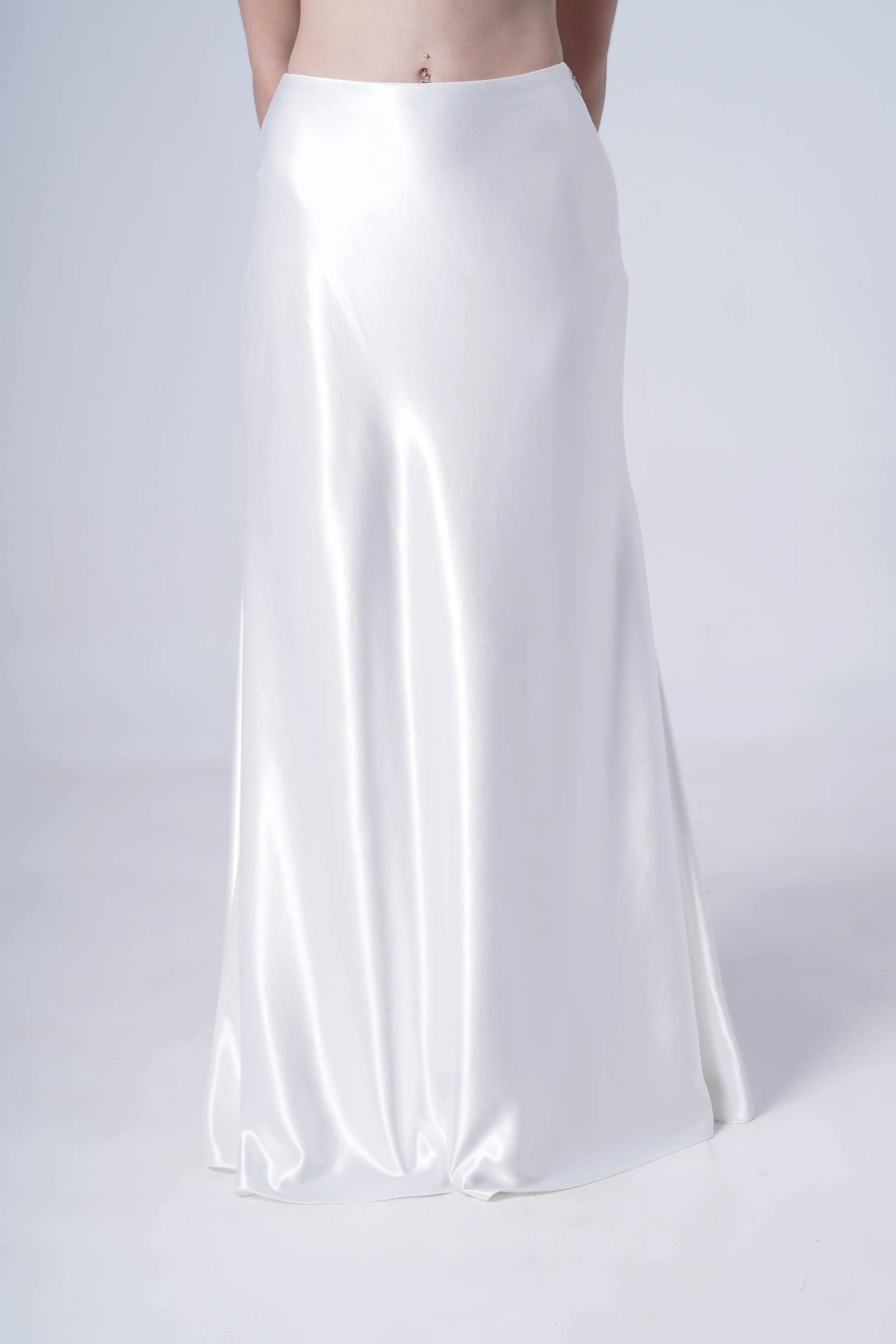 A person models the TH&TH Topaz Bridal Skirt in Ivory Hammered Satin, standing against a plain light background. The long, bias-cut skirt subtly flares, with only the lower torso and skirt visible as their arms rest behind their back.