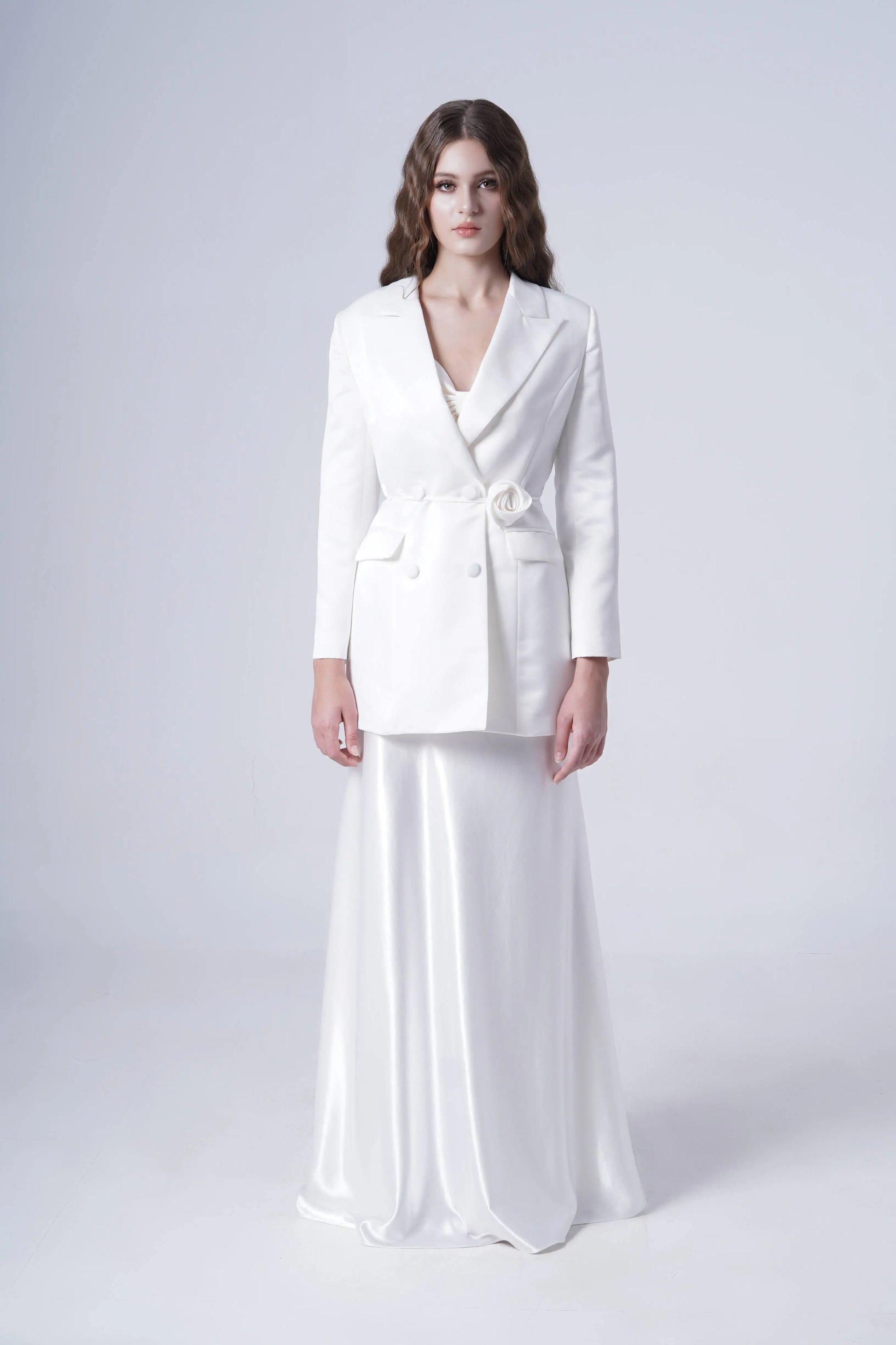 A woman with long wavy hair stands against a plain background, wearing the TH&TH Topaz Bridal Skirt in Ivory Hammered Satin paired with a stylish white blazer featuring a floral detail at the waist.