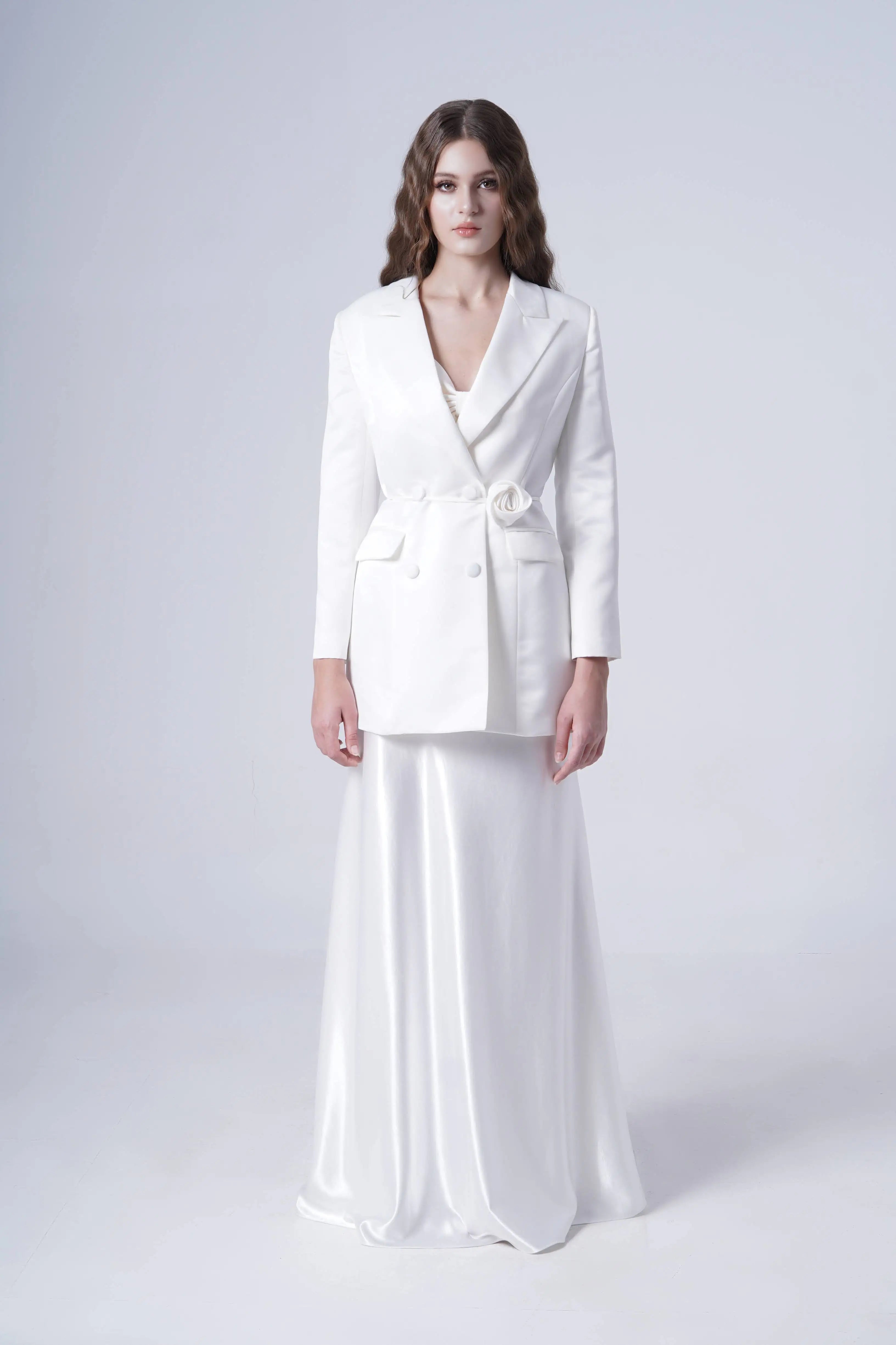 A woman with long wavy hair stands against a plain background, wearing the TH&TH Topaz Bridal Skirt in Ivory Hammered Satin paired with a stylish white blazer featuring a floral detail at the waist.