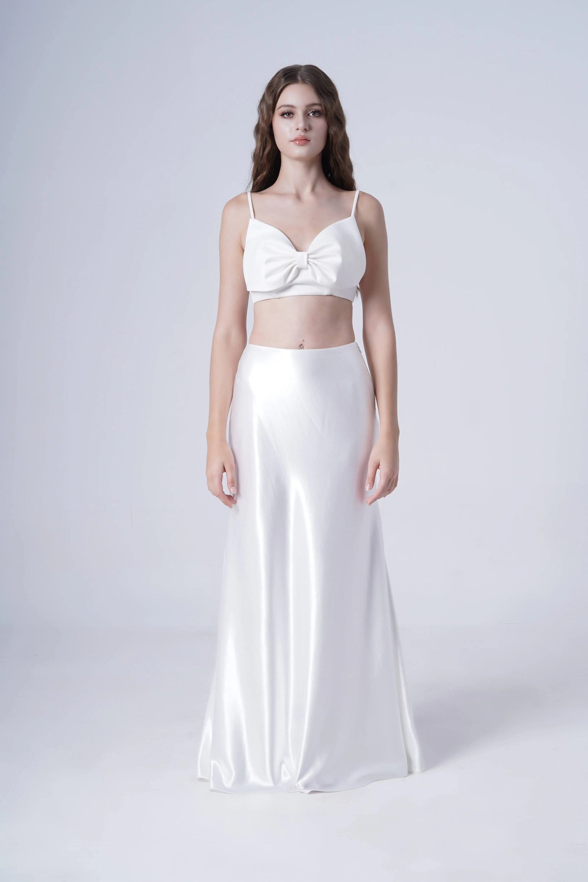 A woman with long, wavy brown hair and a neutral expression wears the TH&TH Topaz Bridal Skirt in Ivory Hammered Satin with a matching white satin crop top featuring a large front knot, standing against a plain light background.