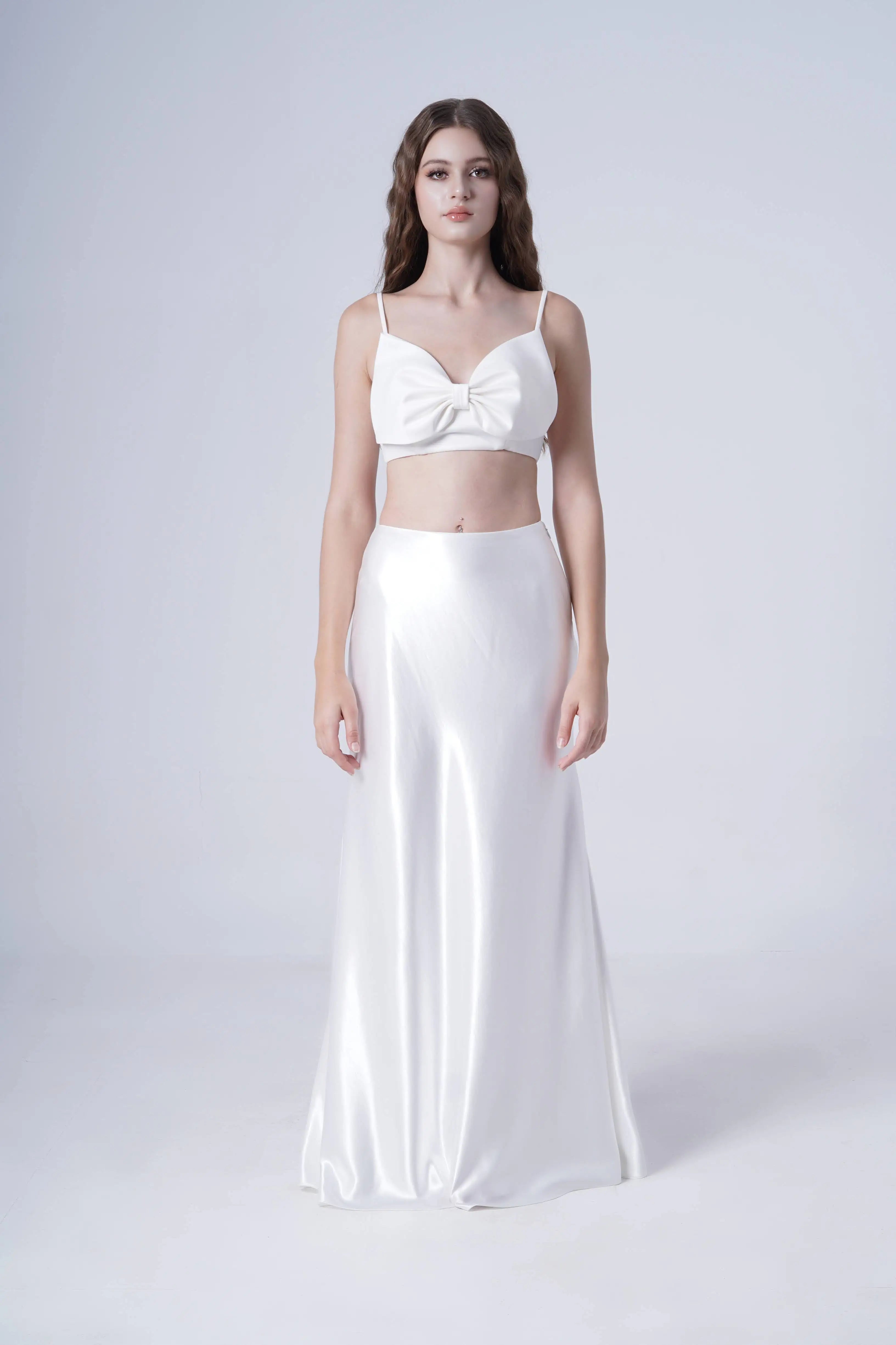 A woman with long, wavy brown hair and a neutral expression wears the TH&TH Topaz Bridal Skirt in Ivory Hammered Satin with a matching white satin crop top featuring a large front knot, standing against a plain light background.