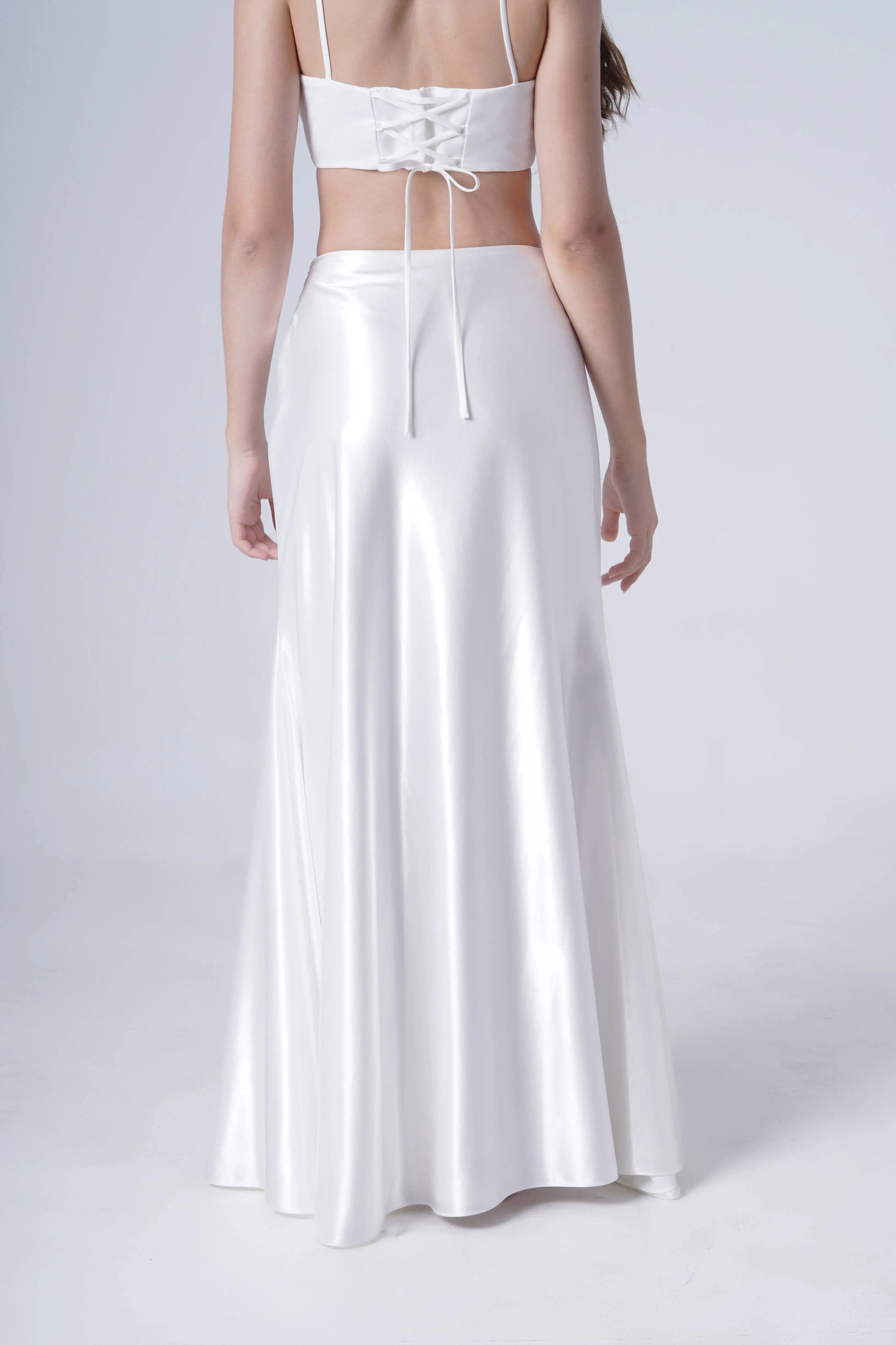 A woman is seen from the back in a light setting, wearing the TH&TH Topaz Bridal Skirt in Ivory Hammered Satin with a matching lace-up crop top. The two-piece outfit features a shiny finish and a flowing, bias-cut skirt.