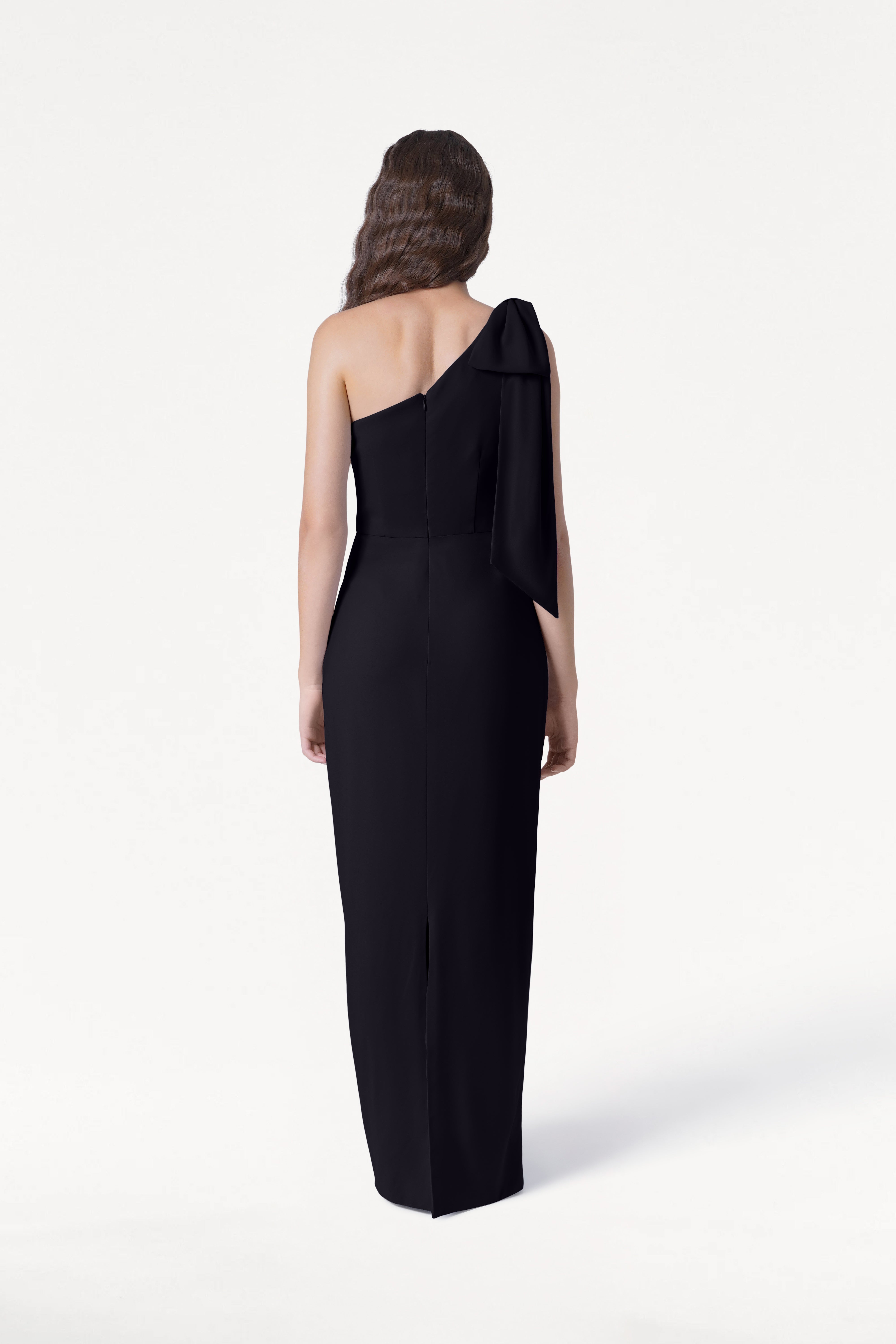 A woman with long wavy hair models the size-inclusive, floor-length Valentina Bridesmaid Dress in Black by TH&TH, featuring a one-shoulder bow detail, as she stands with her back to the camera against a plain white background.