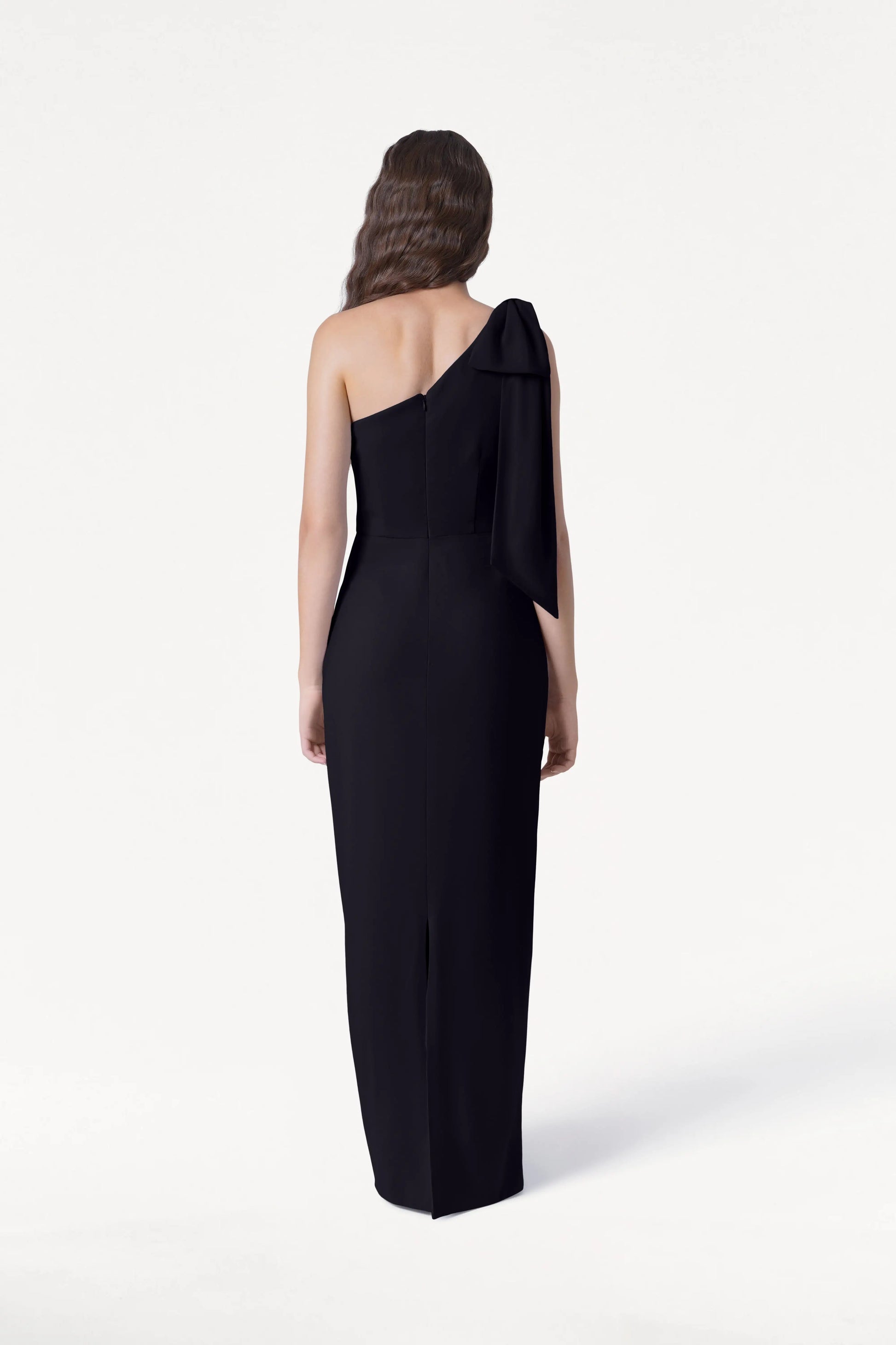 A woman with long wavy hair models the size-inclusive, floor-length Valentina Bridesmaid Dress in Black by TH&TH, featuring a one-shoulder bow detail, as she stands with her back to the camera against a plain white background.