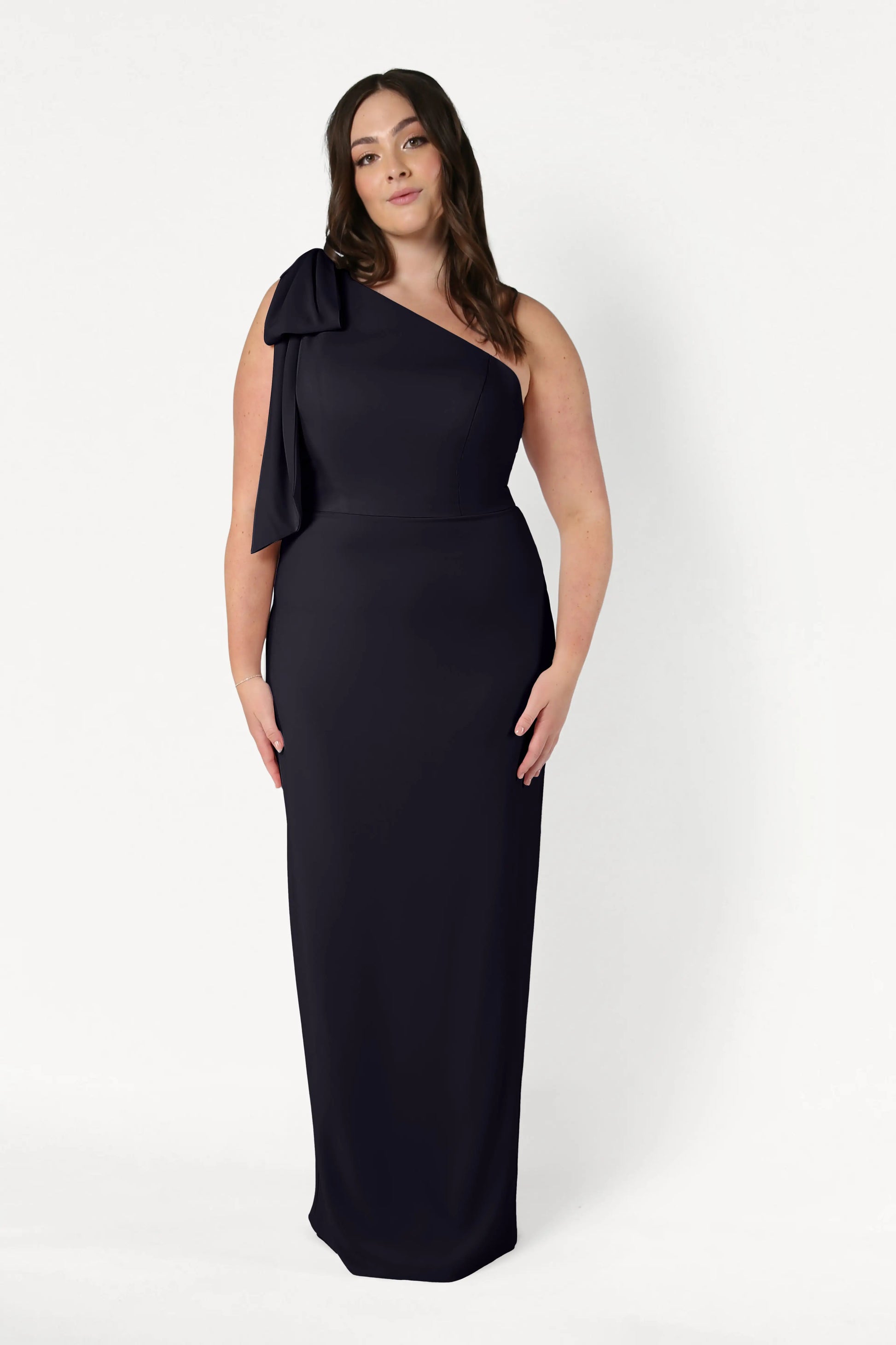 A woman stands against a white background wearing the TH&TH Valentina Bridesmaid Dress in black, a long, one-shoulder style with a bow detail. Her brown hair is styled in loose waves and she has a neutral expression.
