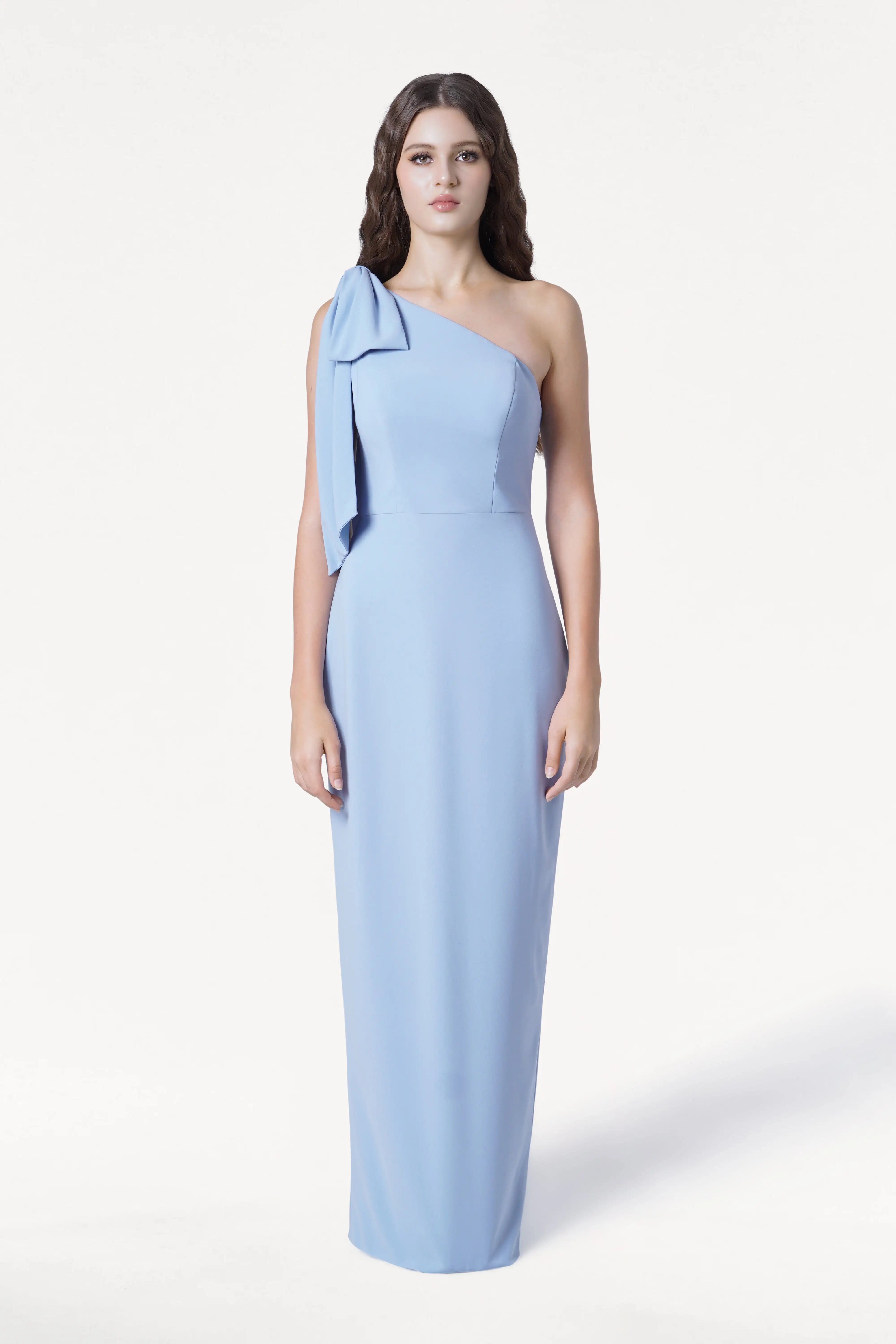 A woman with long, wavy brown hair faces forward in the Valentina Bridesmaid Dress by TH&TH, a modern, dusty blue one-shoulder crepe gown with a left-shoulder bow. She stands against a plain white background with a neutral expression.