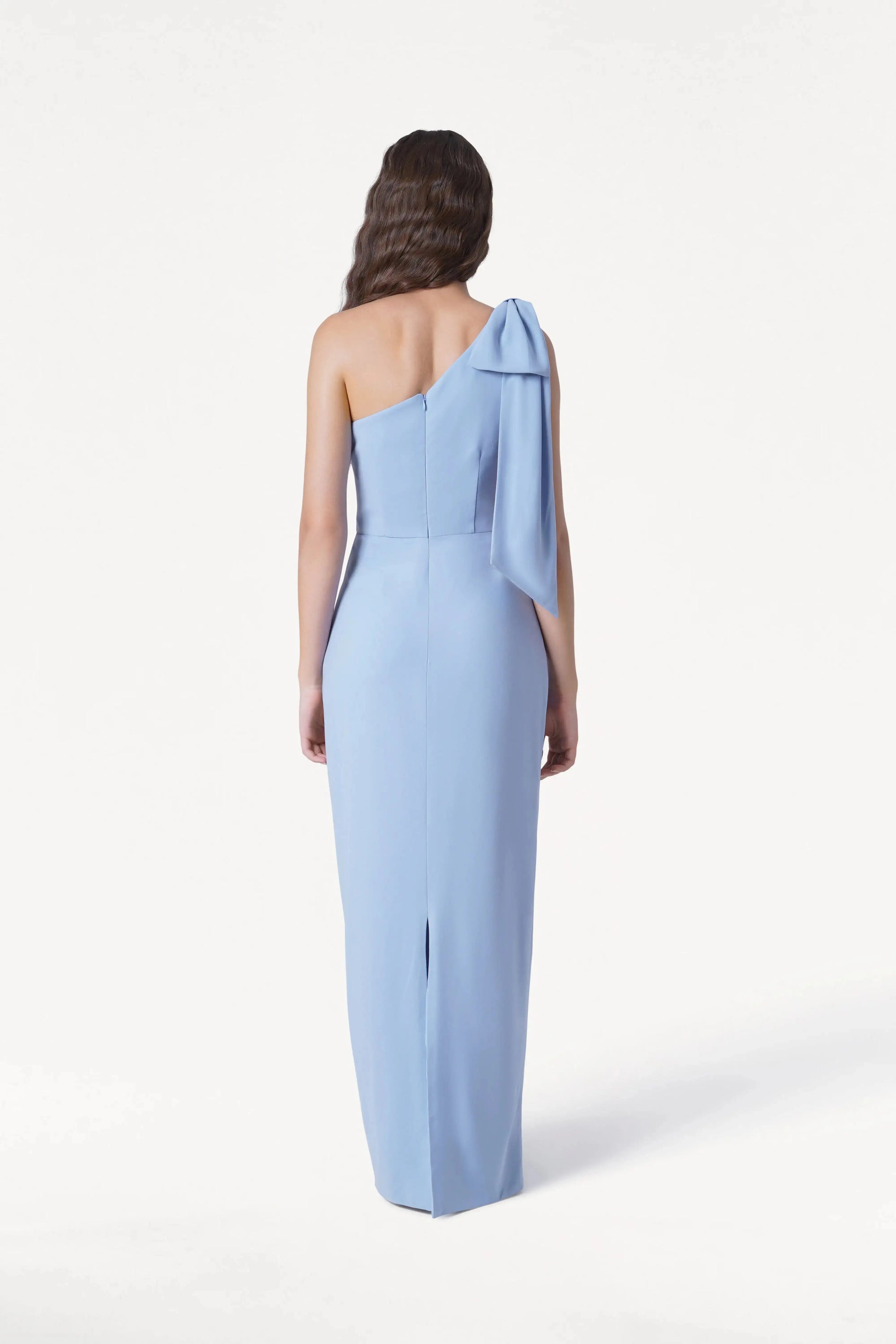 A woman stands facing away, wearing the TH&TH Valentina Bridesmaid Dress in Dusty Blue—a floor-length, one-shoulder crepe gown with a bow on the shoulder. Her long wavy hair flows down her back against a plain white background.